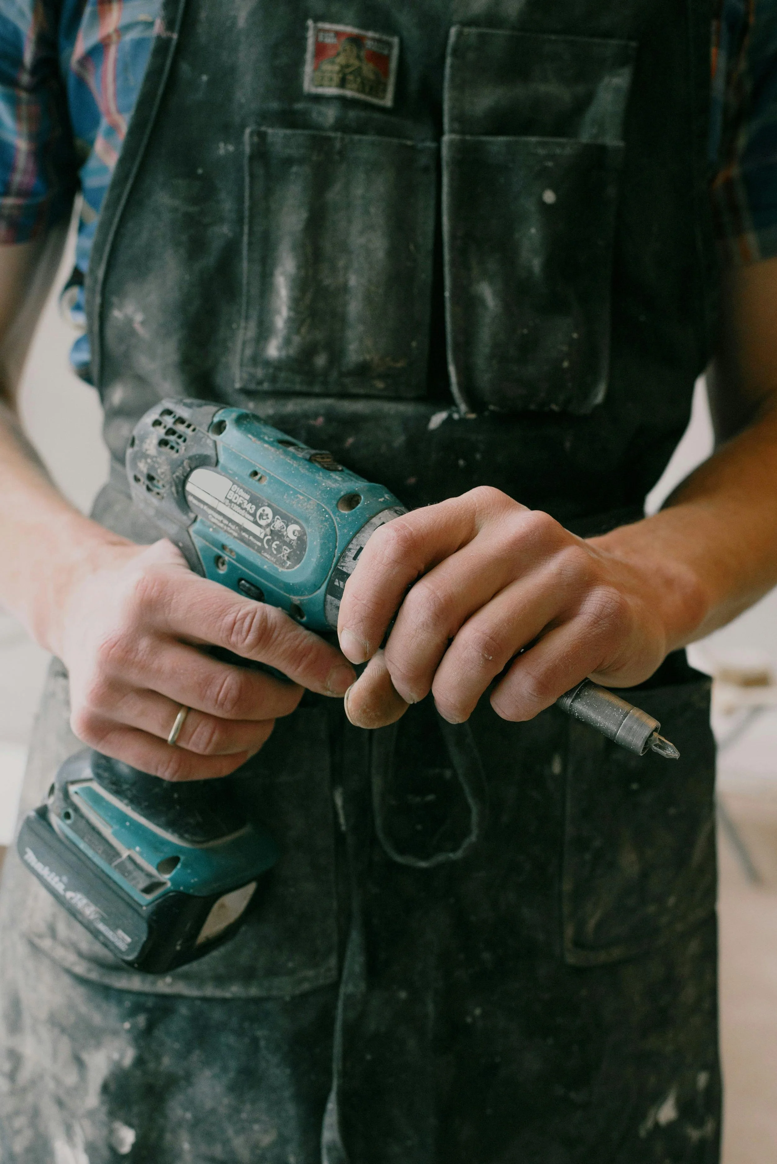 A person in a black work apron holding a cordless electric drill, with a drill bit attached, in a workshop or construction setting.