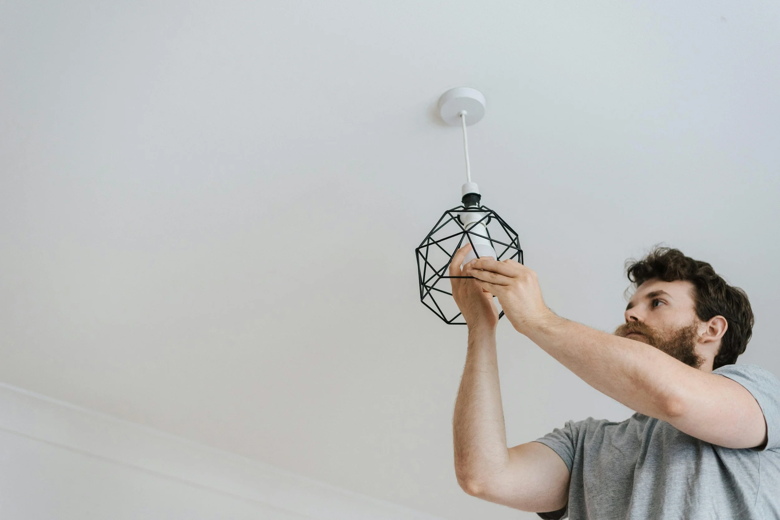 A man installing a modern geometric pendant light fixture on a white ceiling.