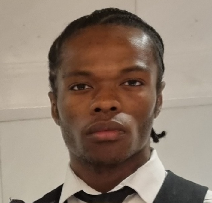 A young man with dark skin and braided hair, wearing a white shirt and a black tie, looking directly at the camera with a neutral expression.