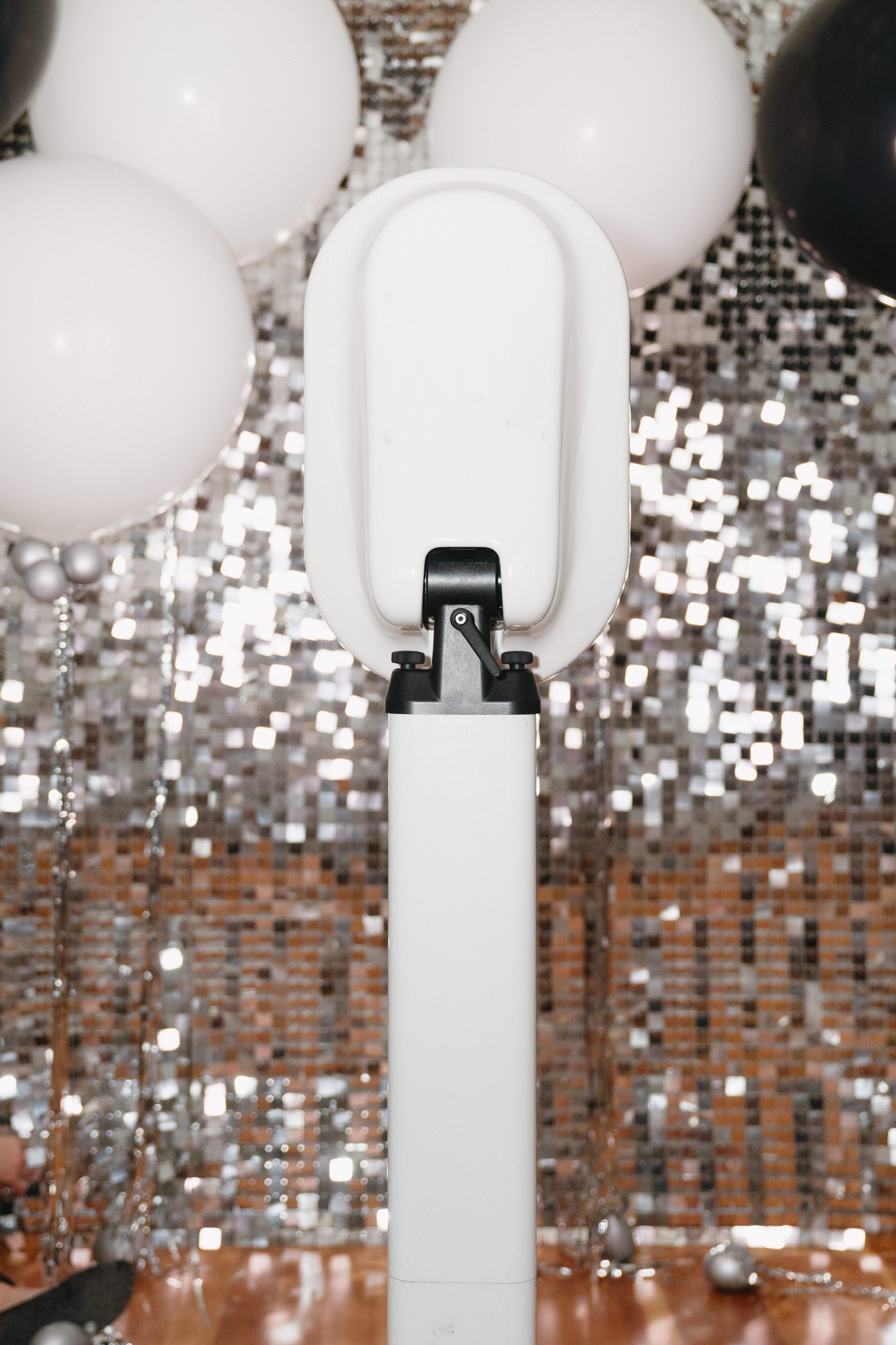 A white device, possibly an air purifier or similar appliance, standing on a white pole, with balloons and metallic decorations in the background on a festive setting, with a brick wall and mirrored tiles.