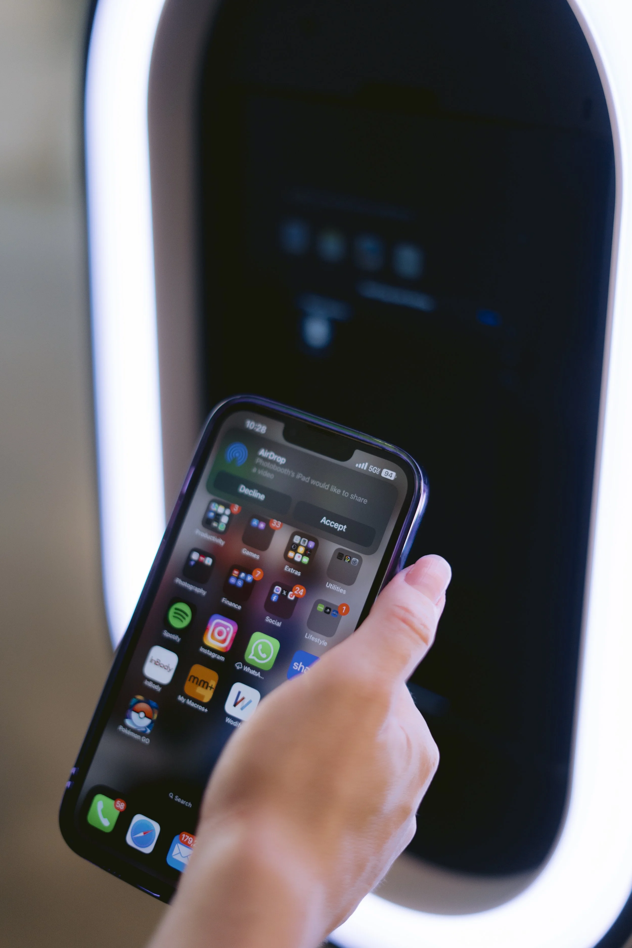 Close-up of a person holding a smartphone displaying app icons and a notification about sharing via AirDrop, with a blurry dark screen in the background.