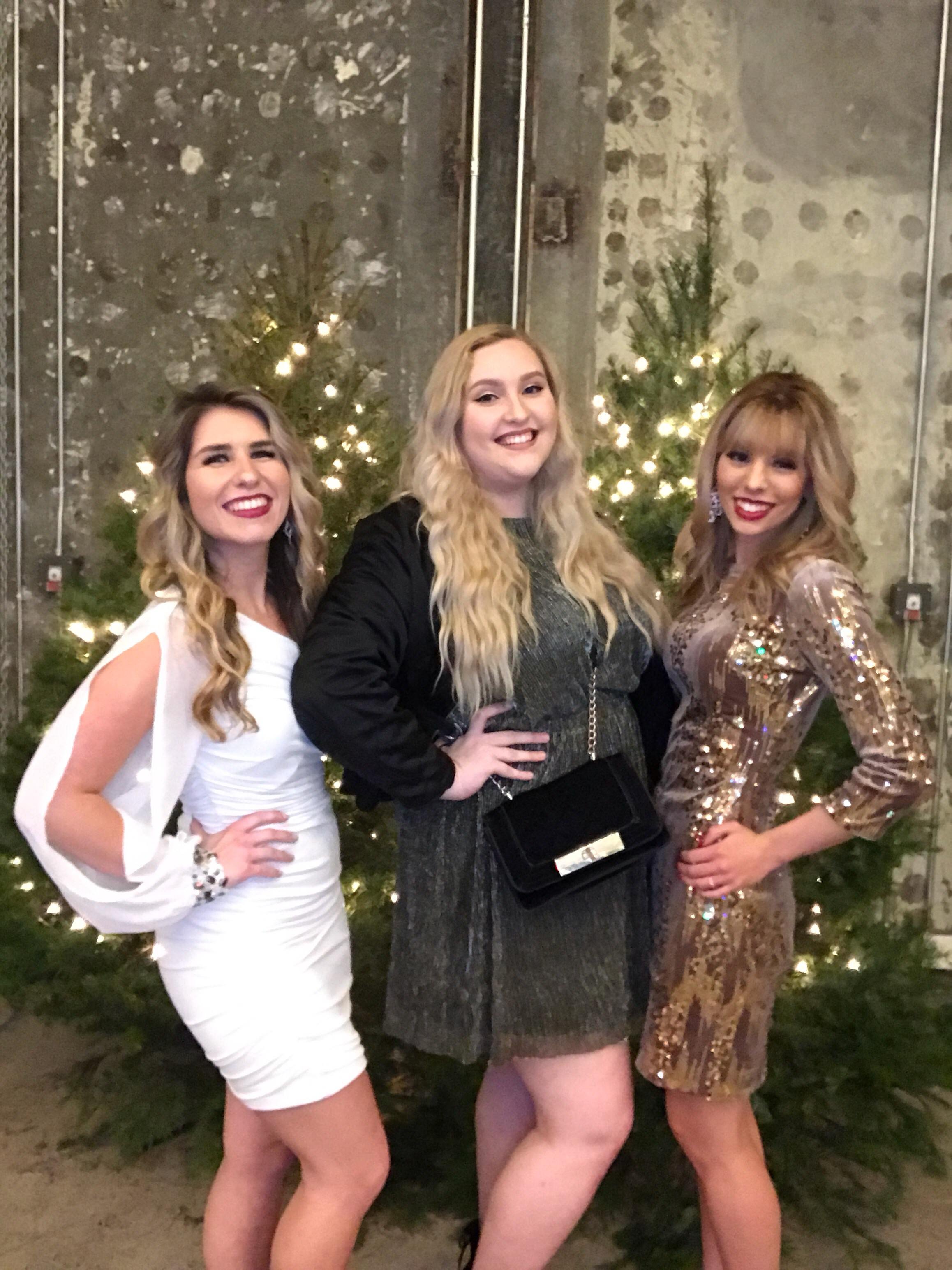 Three women posing in front of decorated Christmas trees with lights, indoors with brick wall background.
