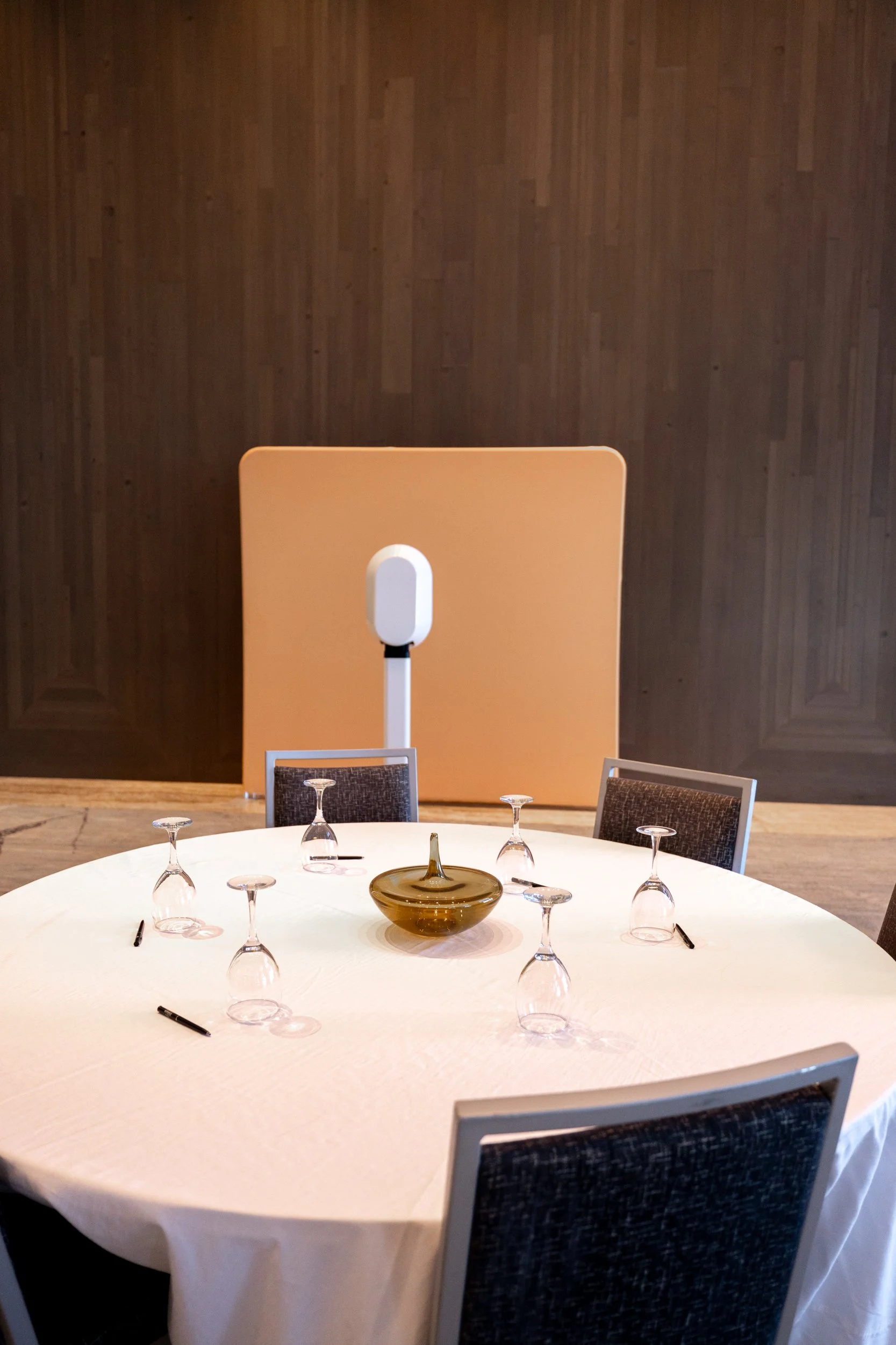 Round dining table covered with a white tablecloth, set with overturned glasses, a central decorative bowl, and black pens. Several chairs are placed around the table. In the background, there is a large beige partition and a dark wooden wall.