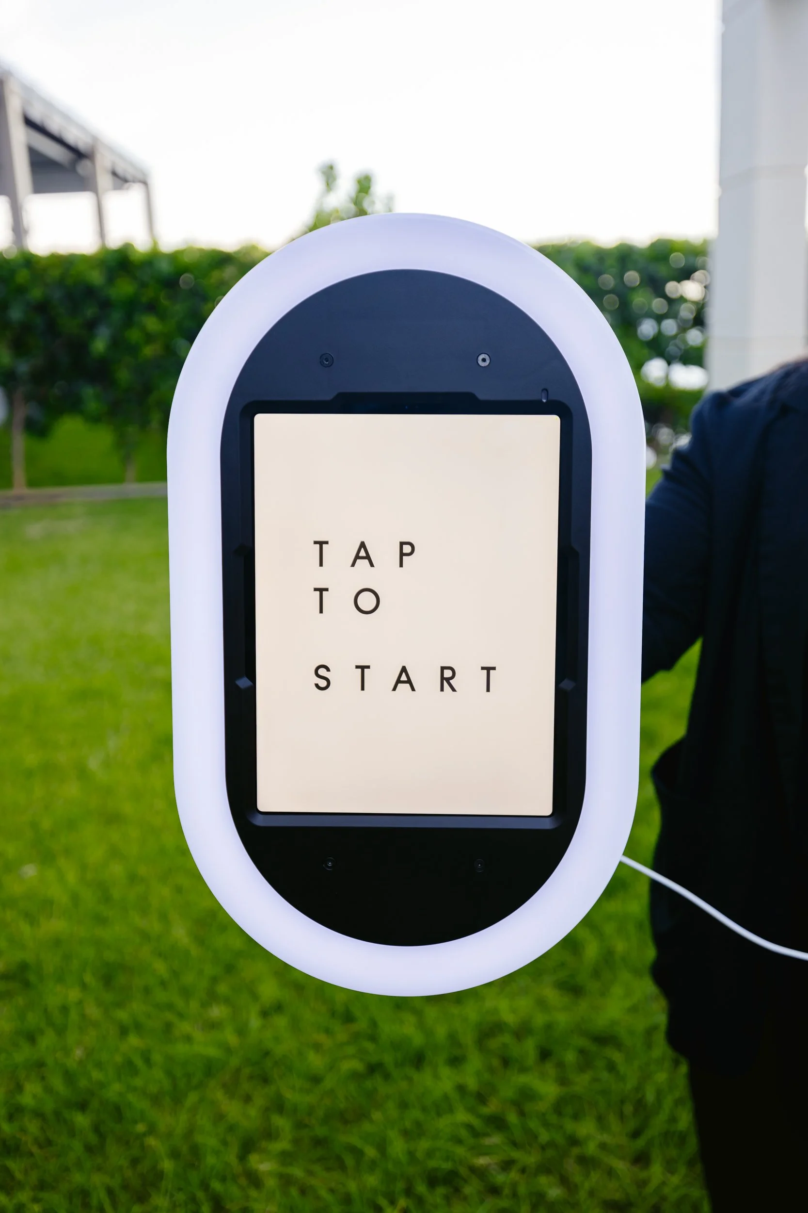 Touchscreen kiosk with a sign that reads 'TAP TO START' outdoors, with grass and trees in the background.