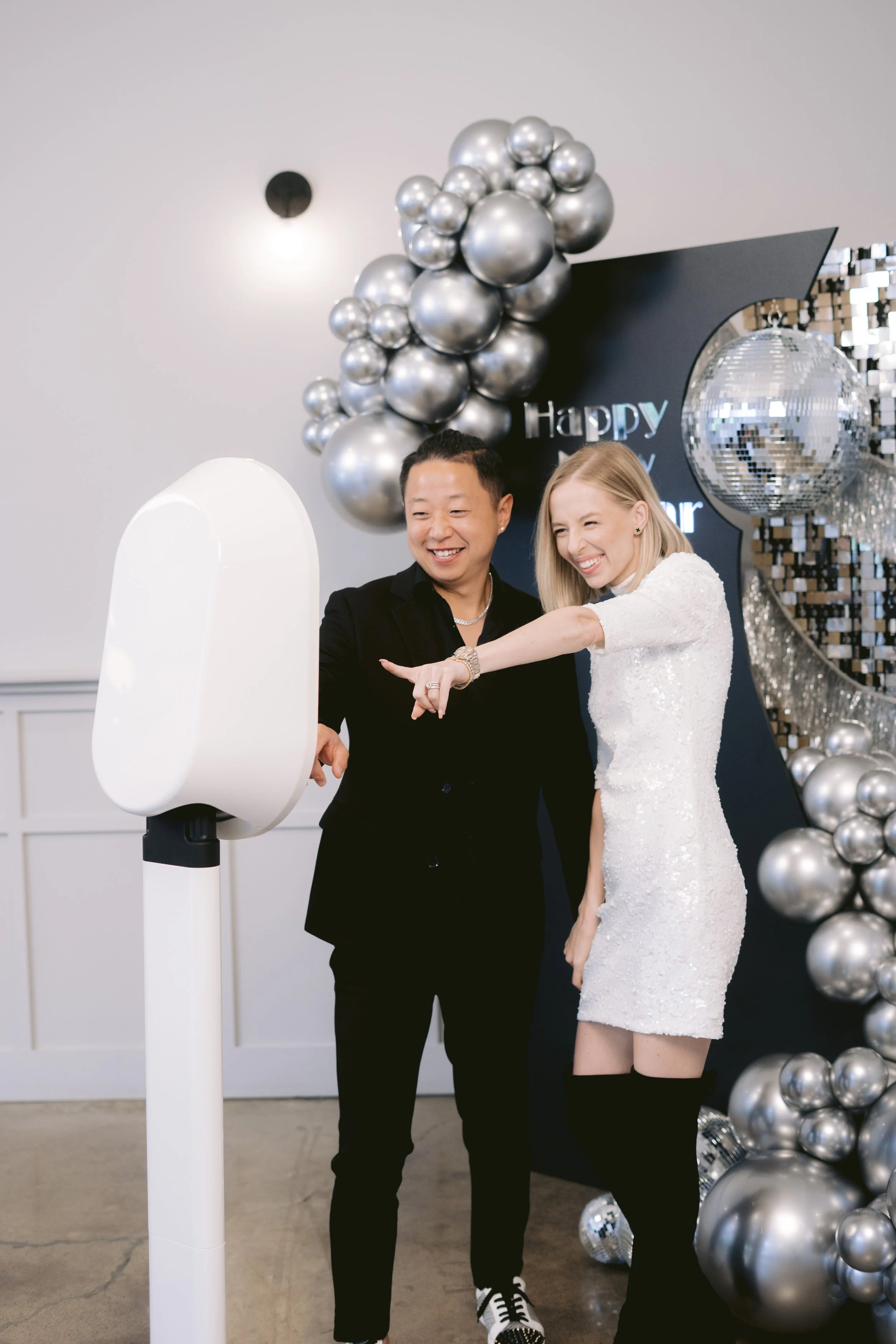 Two smiling people, a man and a woman, seem to be playing a game or participating in a fun activity at a celebration with silver balloon decorations and a disco ball in the background.