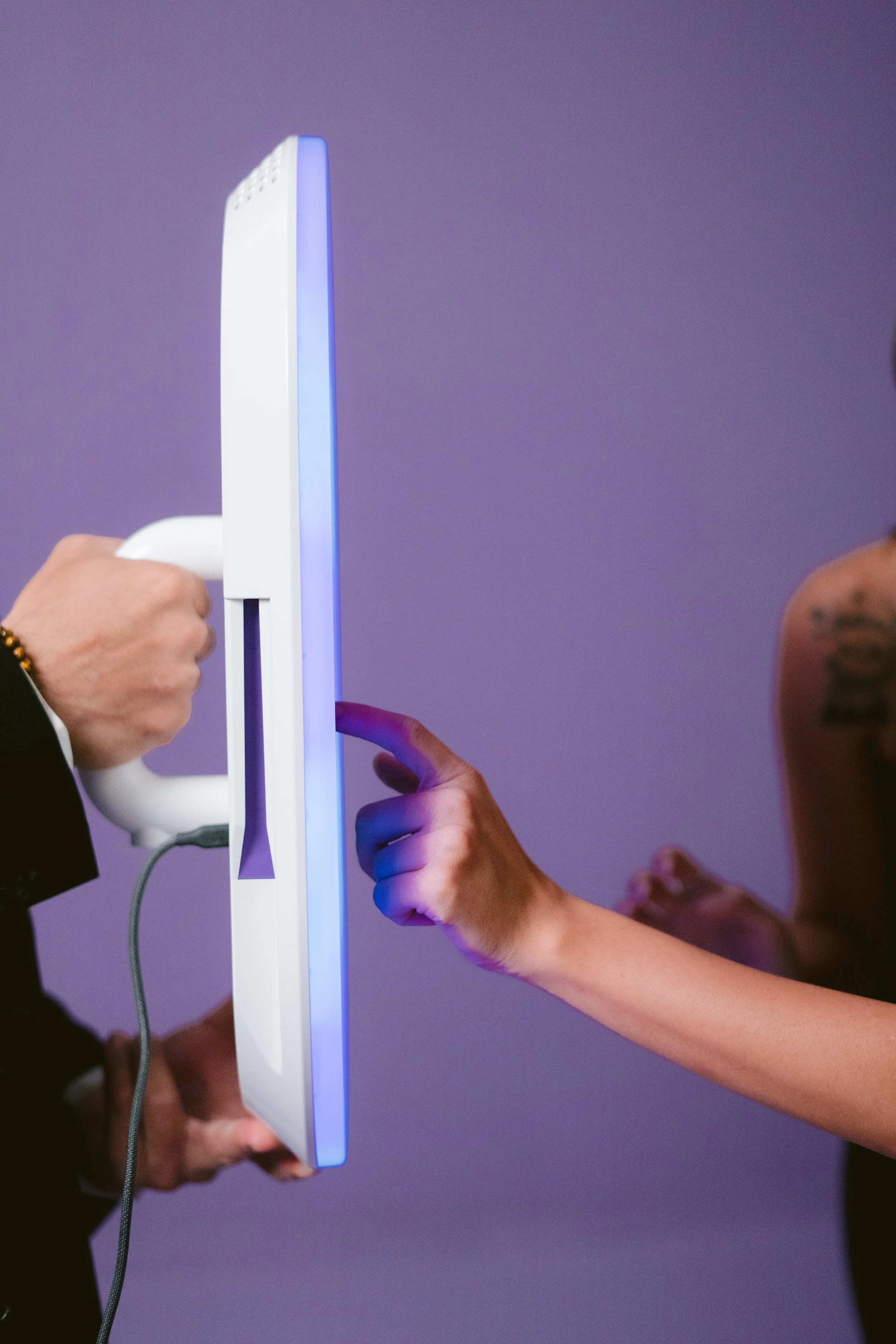 A person is holding a white electronic device with a tall, slim design, illuminated with blue light on the edge. Another person's hand is pointing at the device, and a third person's hand is gripping the device from the side. The background is purple.
