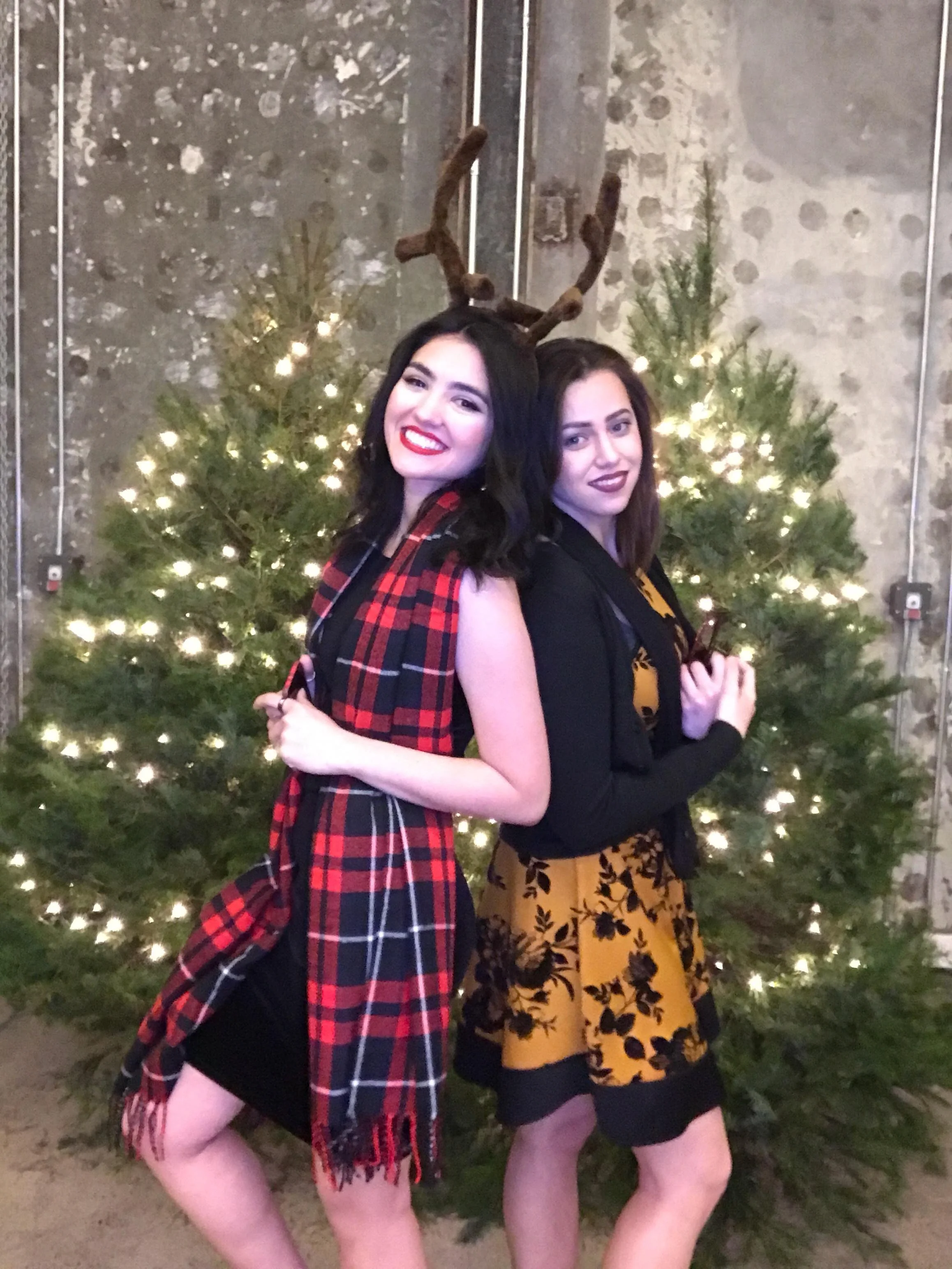 Two women posing in front of a decorated Christmas tree, one wearing a red plaid dress with a reindeer antler headband, and the other wearing a black blazer over a yellow dress with black floral pattern.