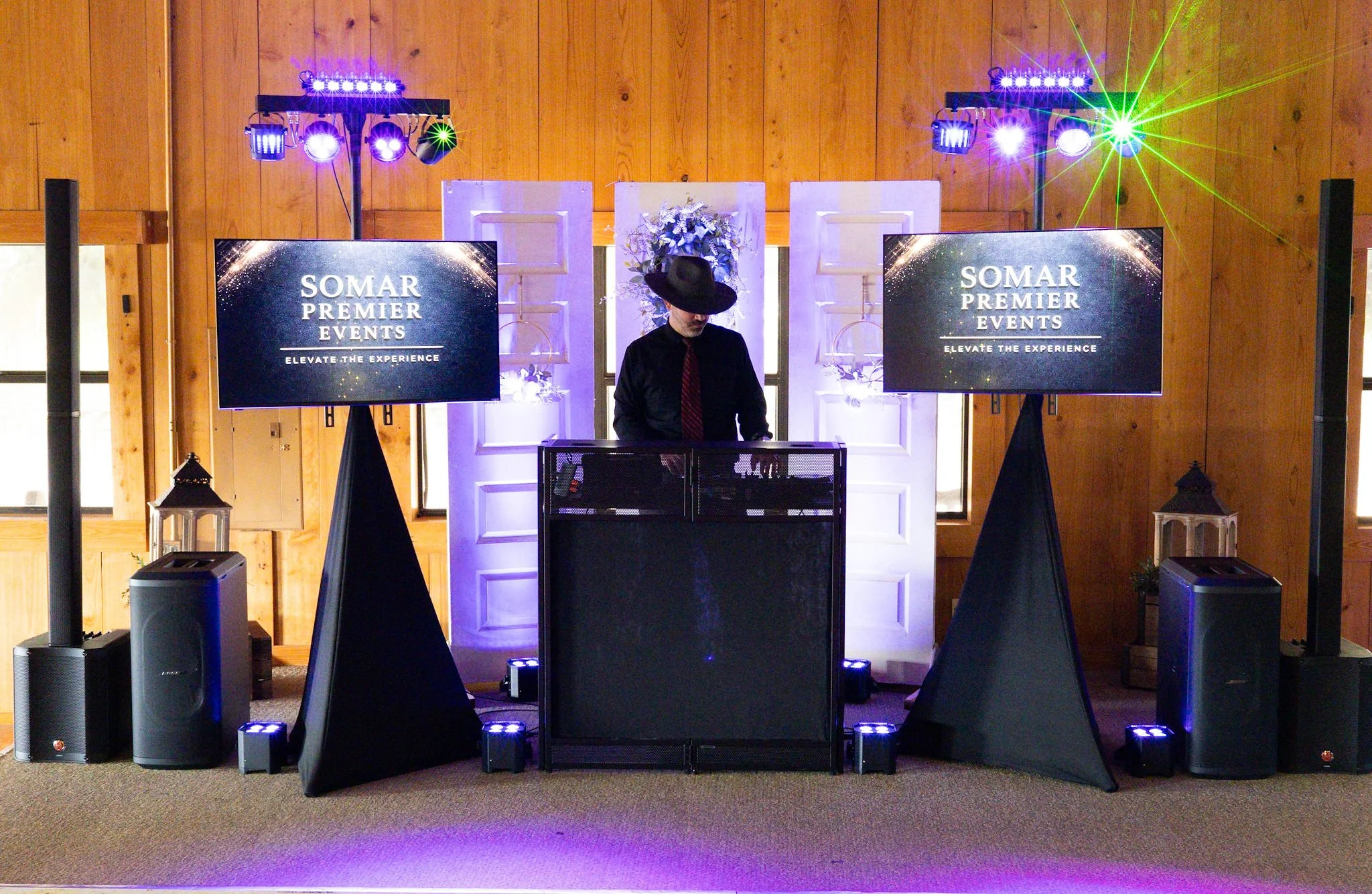 A DJ wearing a black hat and black shirt stands behind a DJ booth at an event. There are two large screens on either side displaying 'SOMAR PREMIER EVENTS - Elevate the Experience.' Colorful lights are shining from the ceiling, creating a vibrant atm