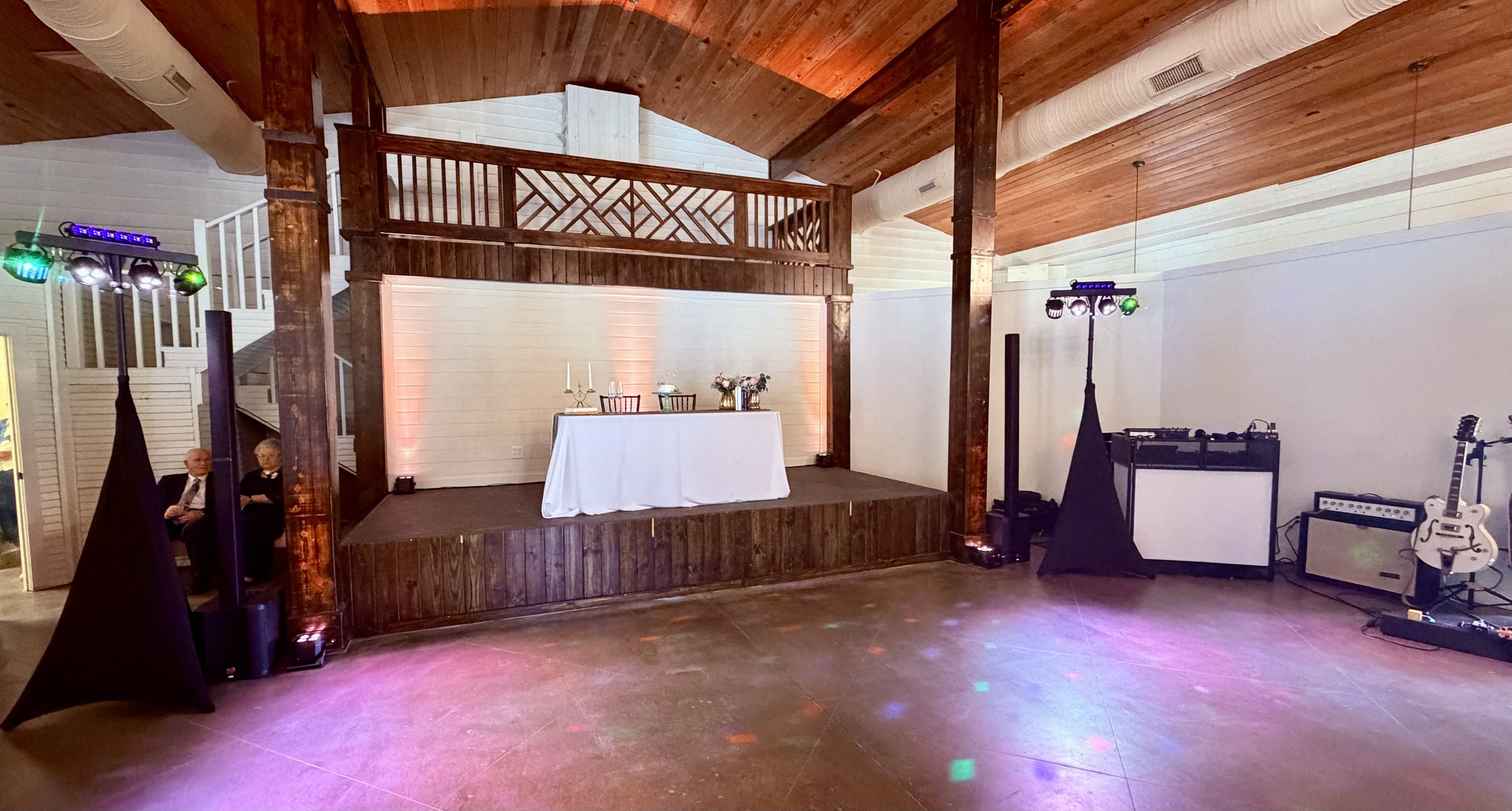 An indoor event space with a small stage decorated with candles and flowers, featuring a wooden balcony and white shiplap walls. There are two people sitting to the left, and DJ equipment with colored lighting and a guitar on the right.