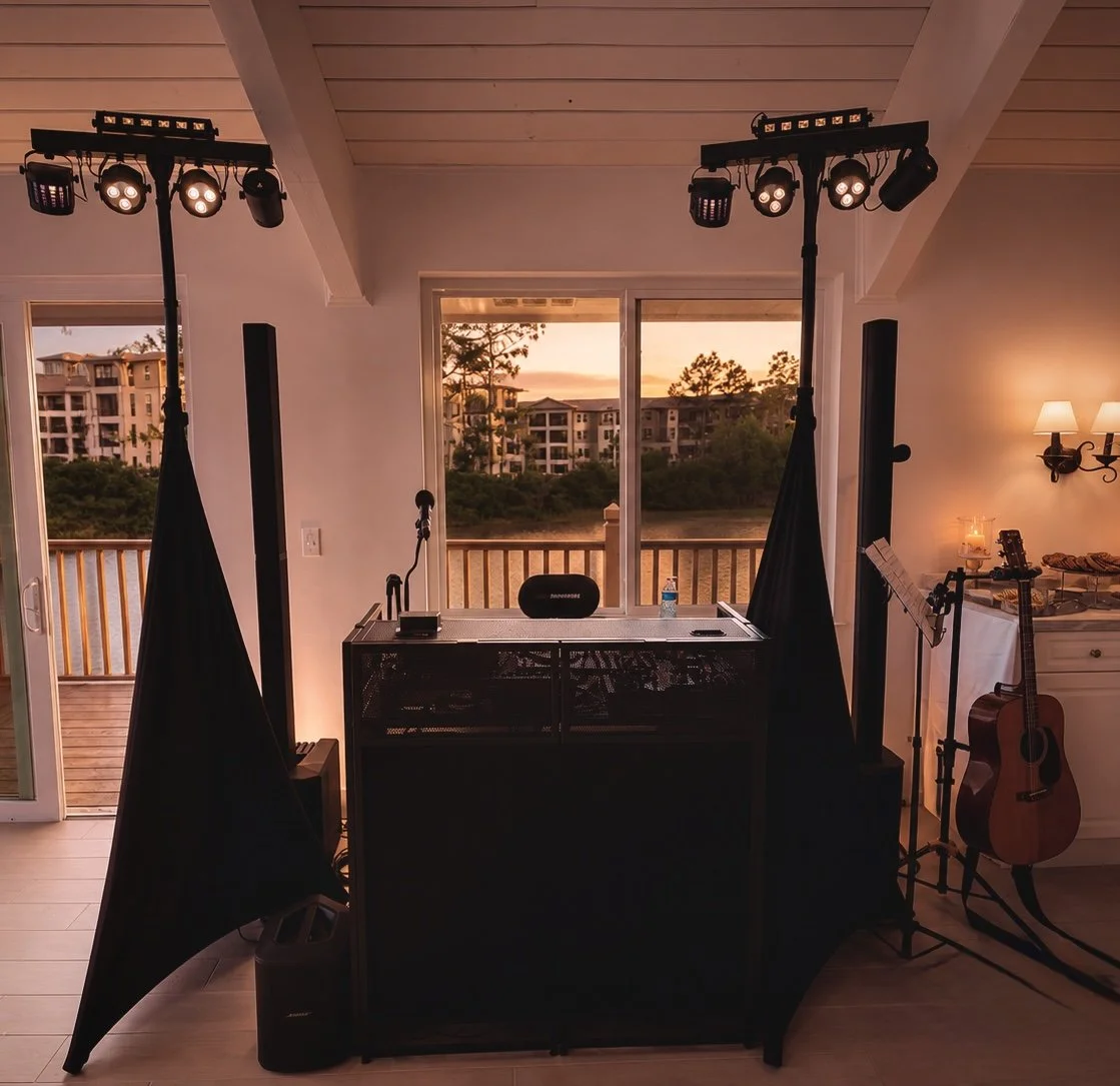 Interior room with DJ setup, including lighting and speaker equipment, a guitar, and a view of an outdoor balcony overlooking trees and multi-story buildings at sunset.
