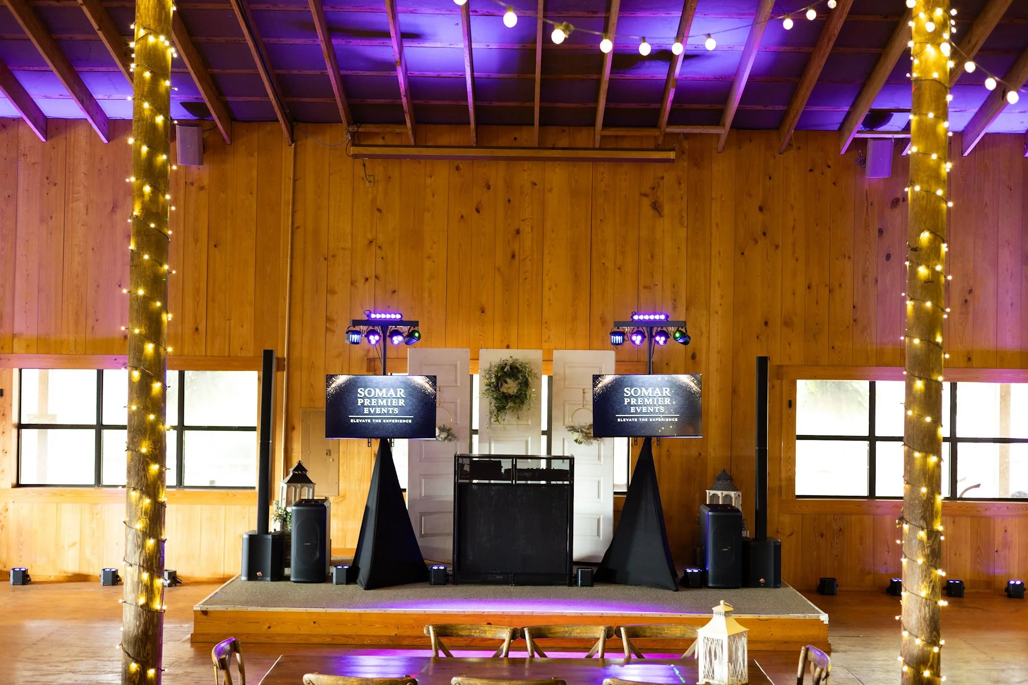 Event stage decorated with wooden walls, fairy lights wrapped around wooden posts, two screens displaying 'SOMAR PREMIER EVENTS', speakers, lighting equipment, a DJ booth, and decorative lanterns in front.