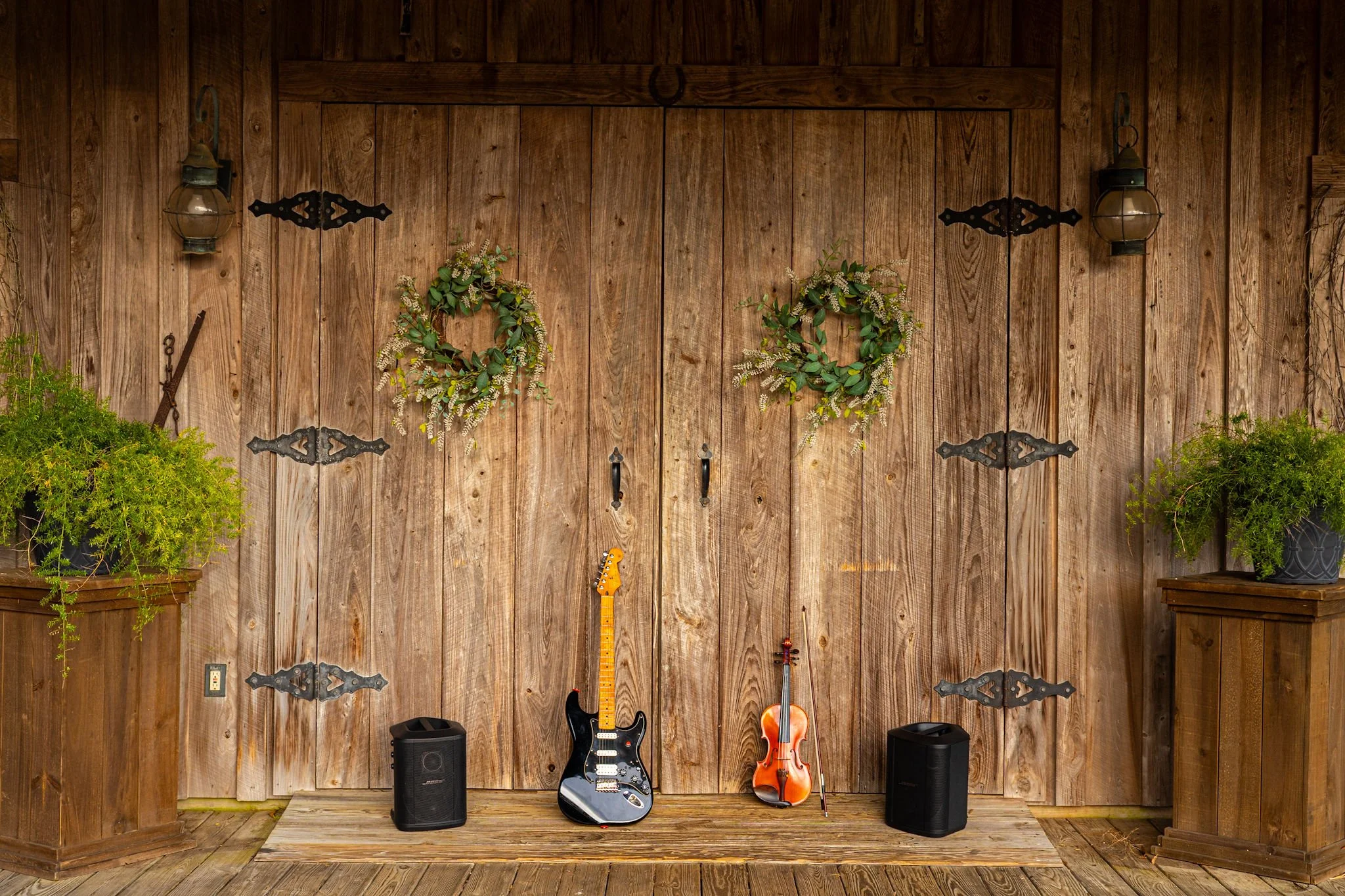 A rustic scene featuring a wooden wall decorated with two green wreaths, plants in planters, and musical instruments, including an electric guitar and a violin with a bow, along with two black speakers.
