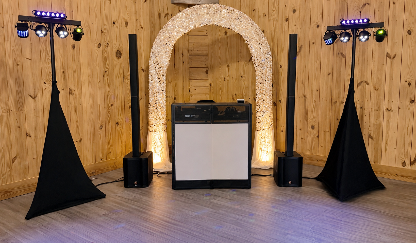 DJ booth setup with speakers and lighting on a wooden floor in front of a wood-paneled wall, featuring a decorated arch backdrop.