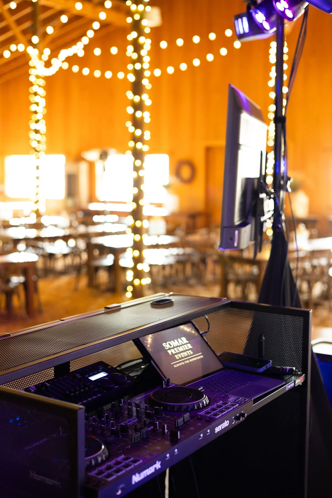 DJ equipment set up with a monitor displaying 'SOMAR PREMIER EVENTS' inside a venue with hanging string lights and wooden walls.