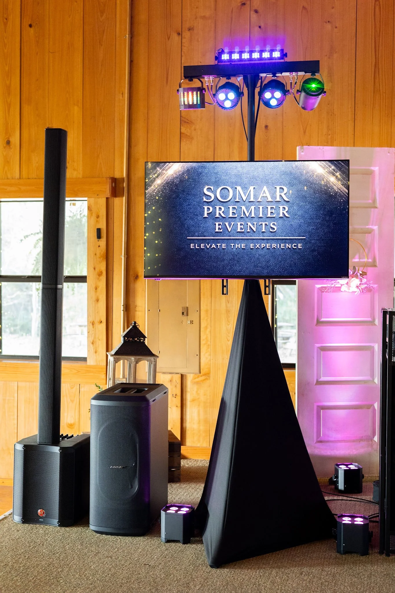 A large flat-screen TV displays 'SOMAR PREMIER EVENTS ELEVATE THE EXPERIENCE.' The TV is on a stand with speakers, lighting equipment, and a pink decorative wall panel in the background, indicating a setup for an event or presentation.
