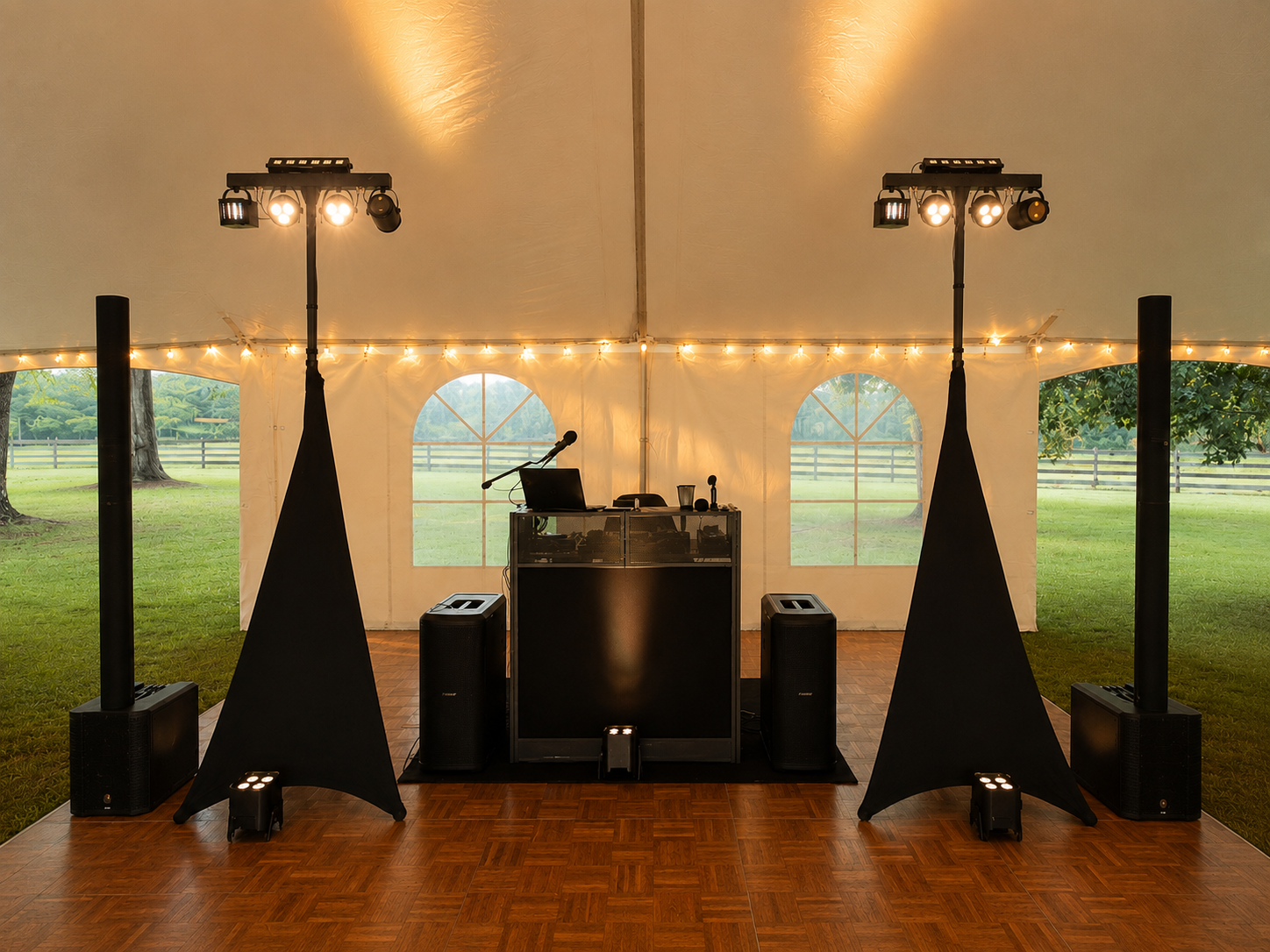 Wedding or event reception setup inside a large tent with a DJ station, speakers, microphones, and stage lights, overlooking a green outdoor area with trees and a fence.