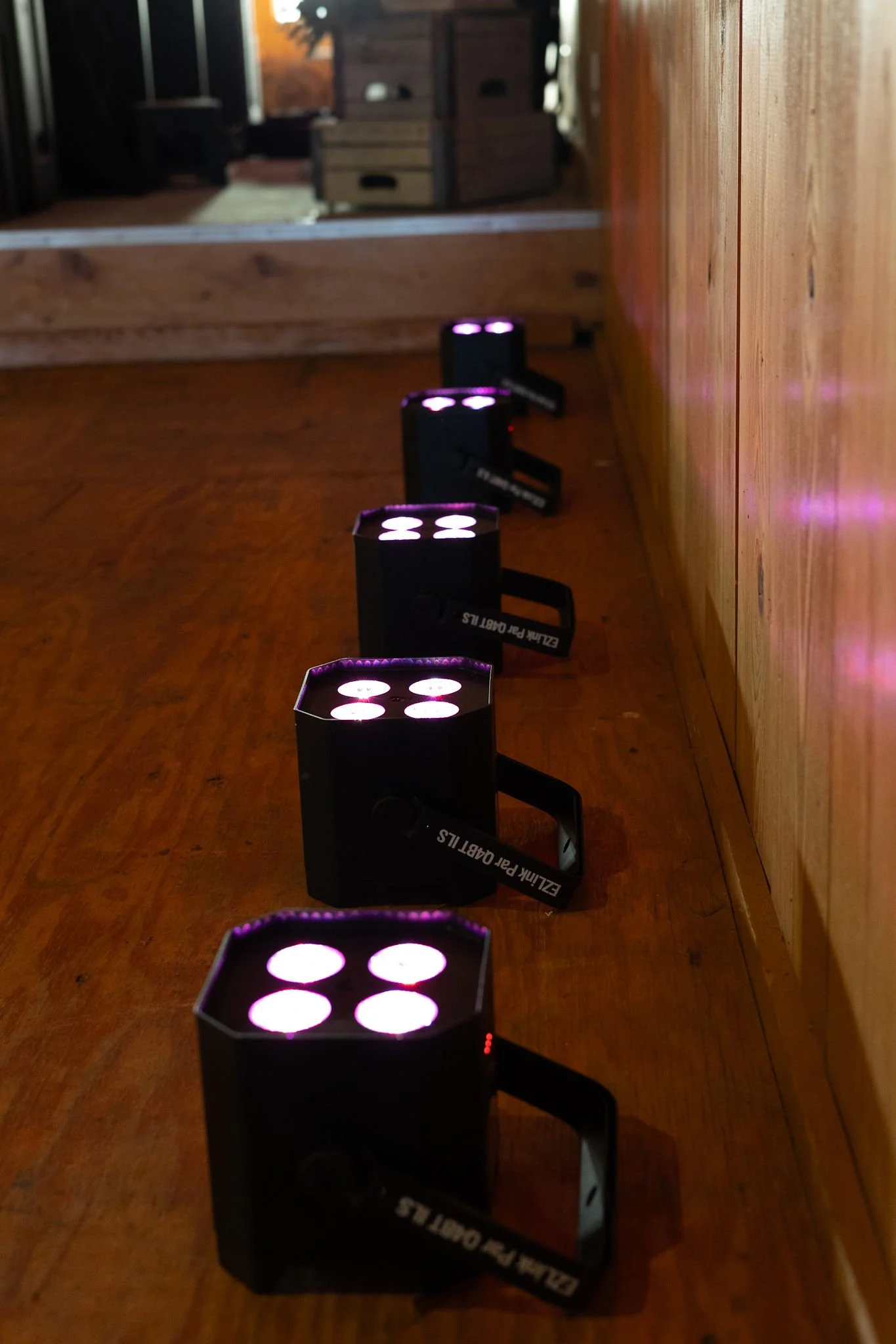 A row of five small black stage lights with four purple LED bulbs each, directed towards a wooden floor and wall.