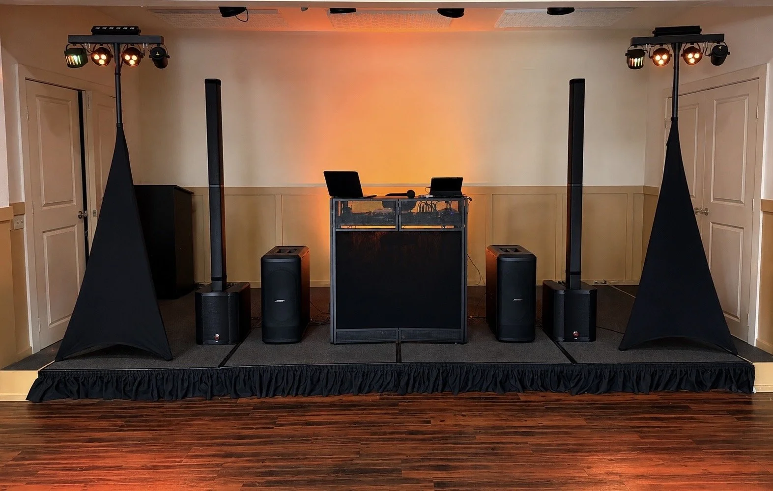 DJ setup on a stage with speakers, lighting, and a table with DJ equipment, in a room with wooden flooring and beige walls.