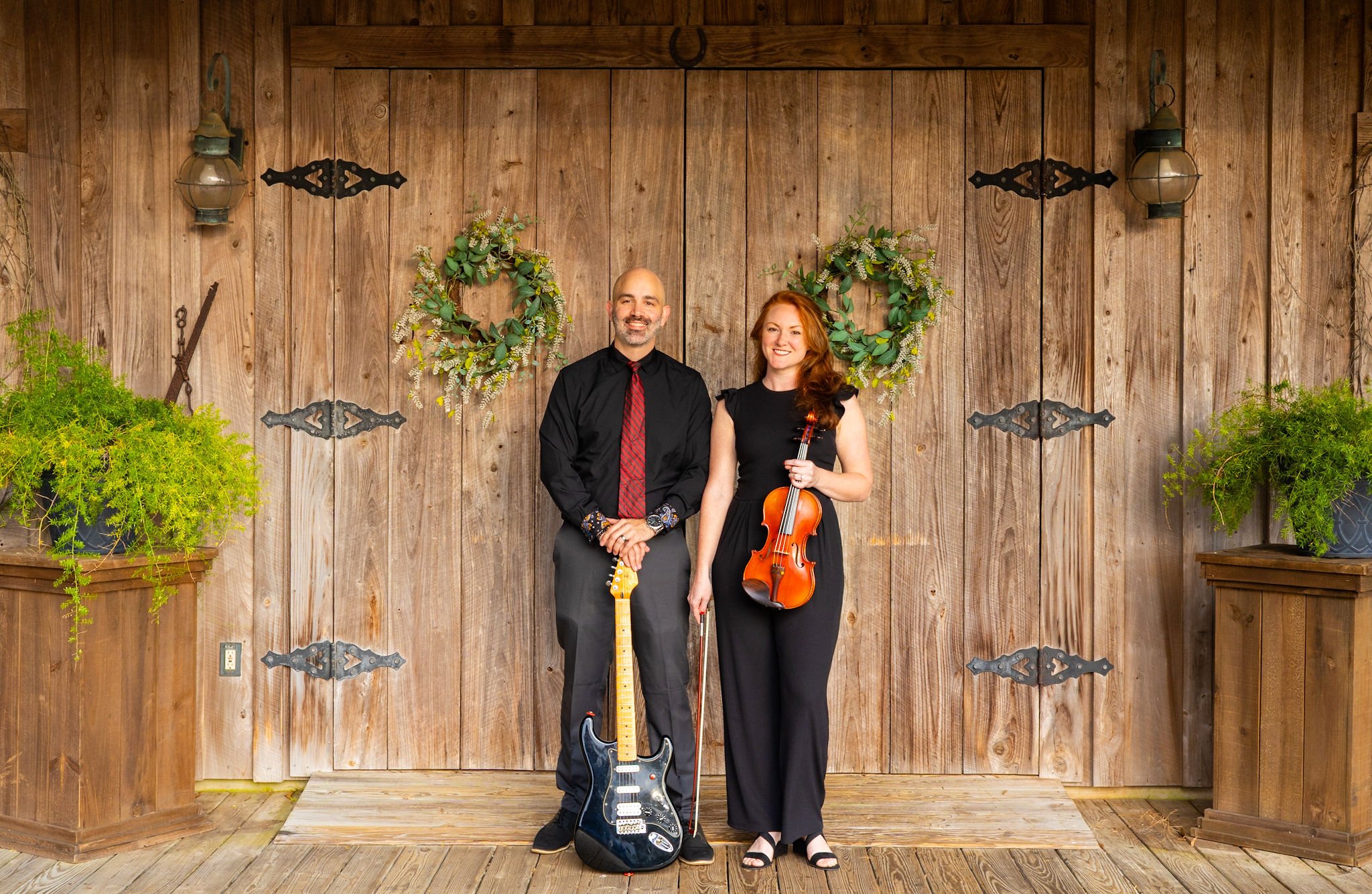 A man and woman standing in front of wooden barn doors decorated with wreaths and lanterns. The man holds an electric guitar and the woman holds a violin.