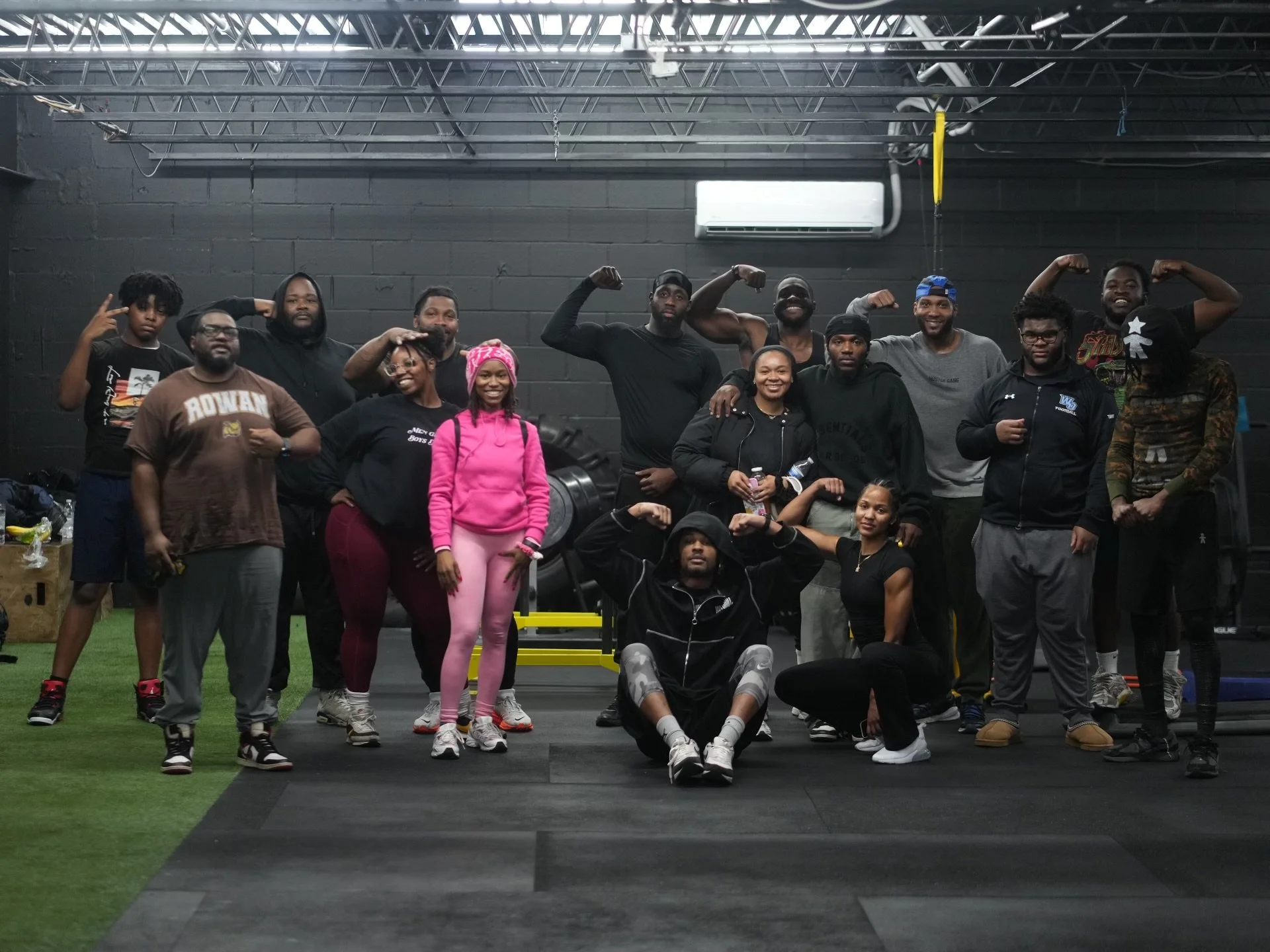 Group of people posing inside a gym or fitness facility, some flexing muscles. Black walls, gym equipment and a green turf area are visible in the background.