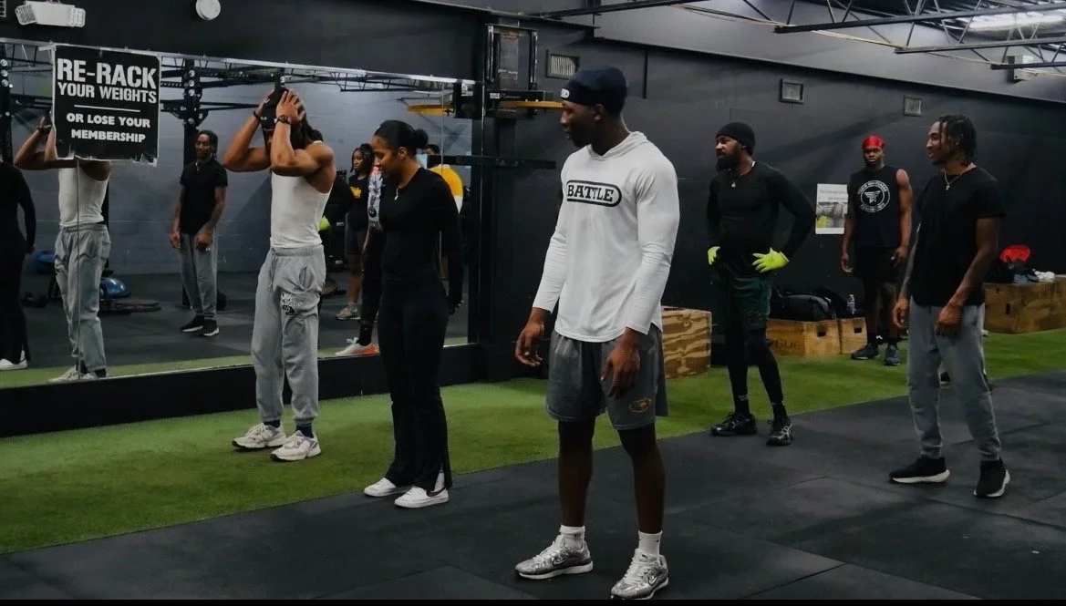 Group of people in a gym, some standing and others exercising, with signage on the wall and workout equipment visible.