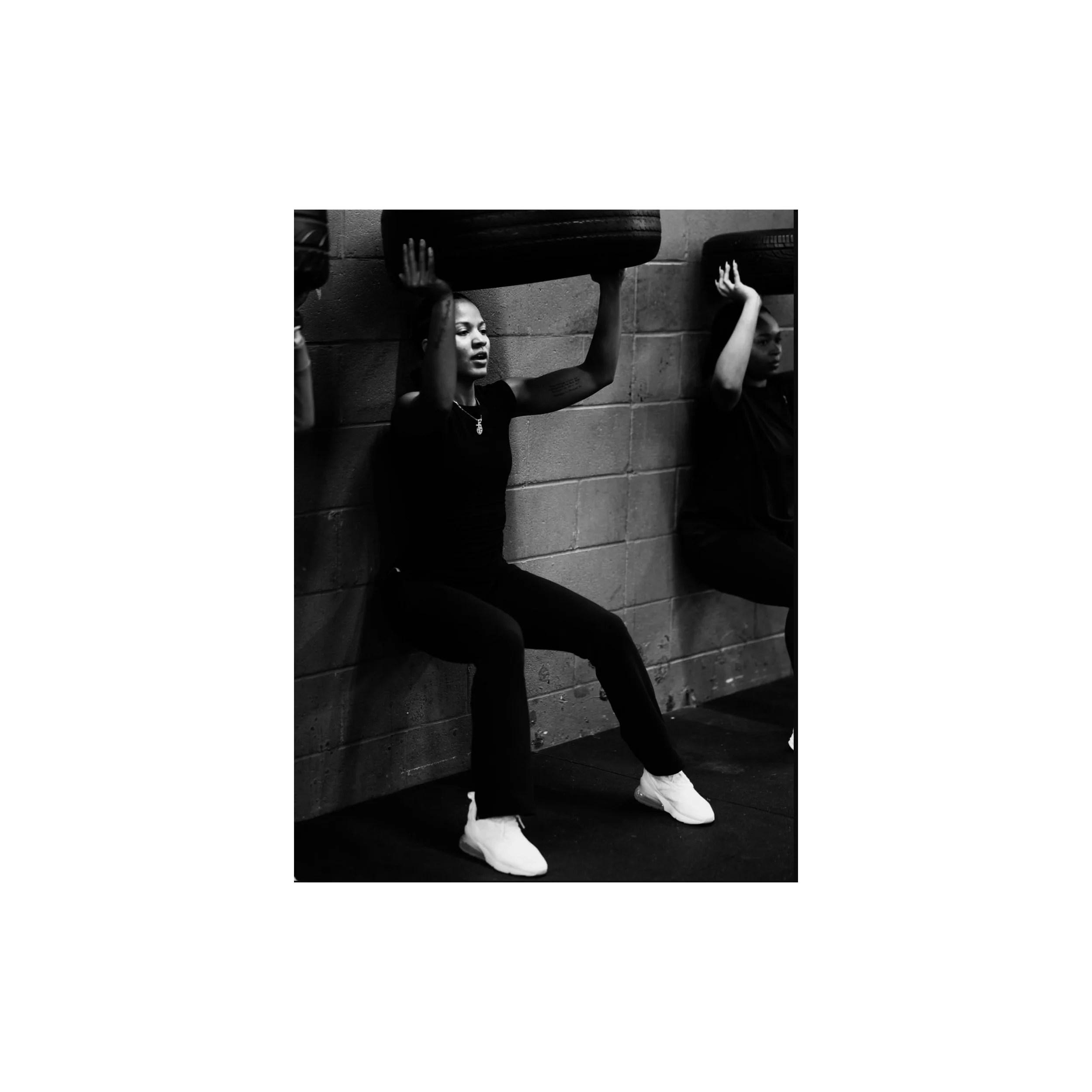 Two women sitting against a brick wall, holding tires overhead. The woman on the left has earrings and a necklace, with her eyes closed. The woman on the right has a short hairstyle, both dressed in black, white sneakers, in a fitness or gym setting.