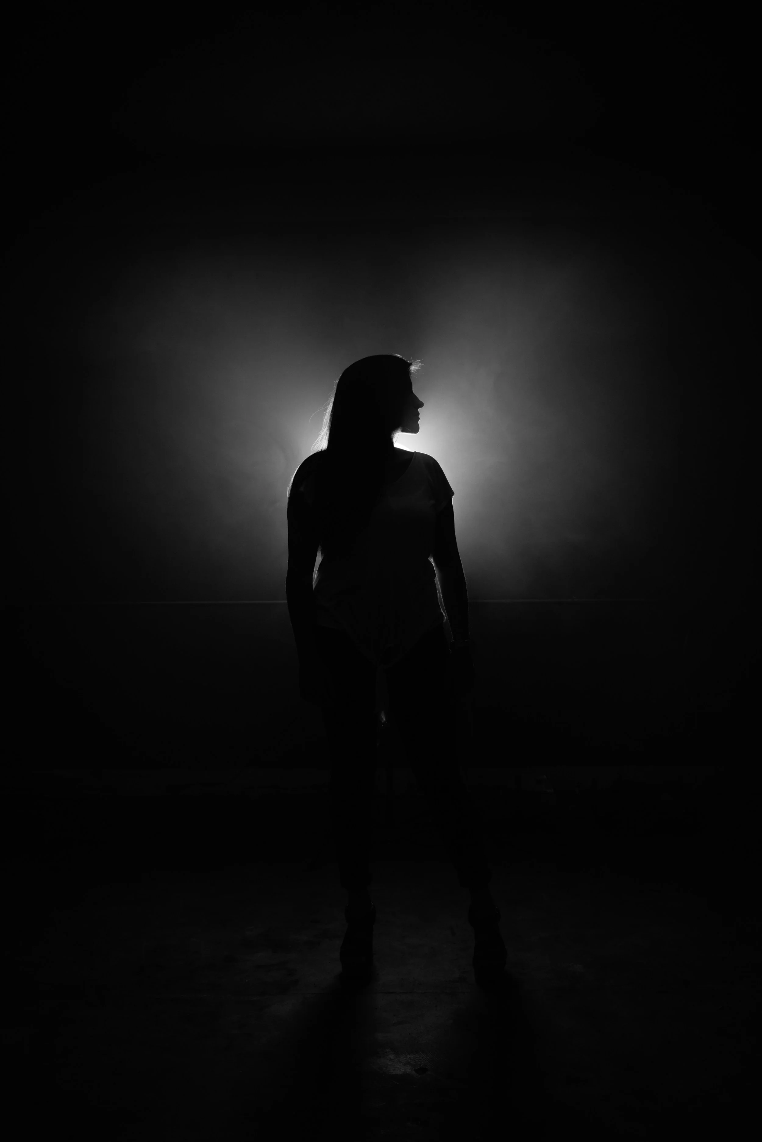 Silhouette of a woman in the darkness with a light in the distance. Her head is turned to the right as if she is looking over her shoulder at you.