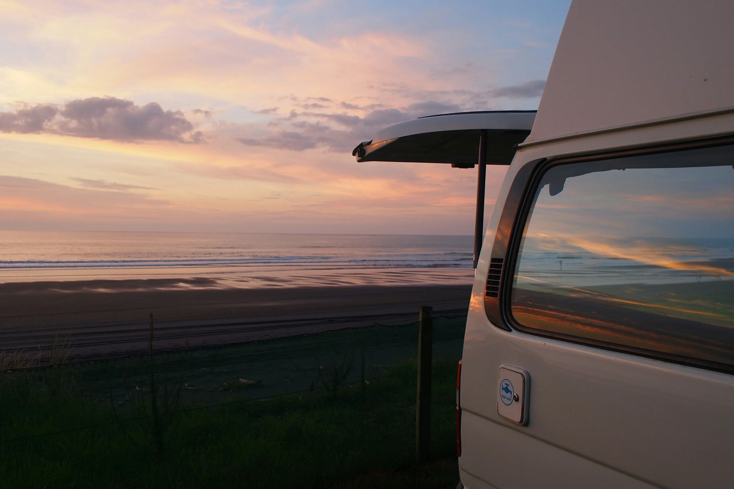 A campervan parked near the beach at sunset, with the ocean and cloudy sky in the background, reflecting in the campervan's window.