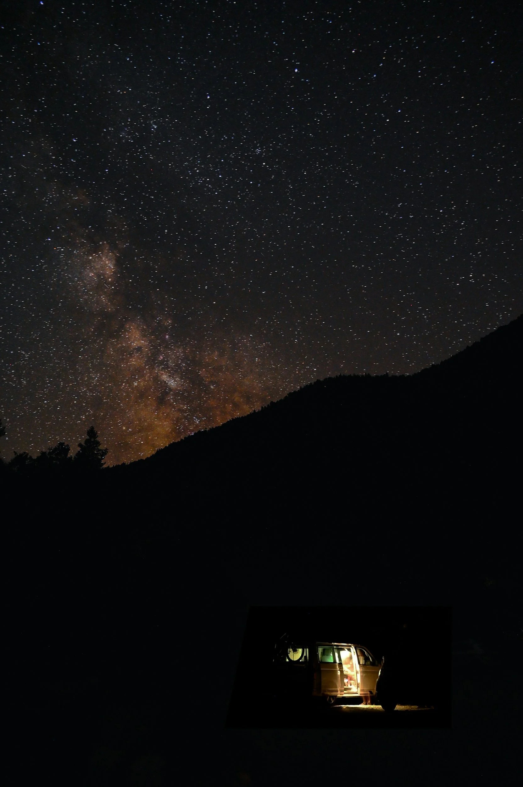 Night sky filled with stars and the Milky Way galaxy, silhouette of a mountain and a van with its sliding door open and interior lights on.