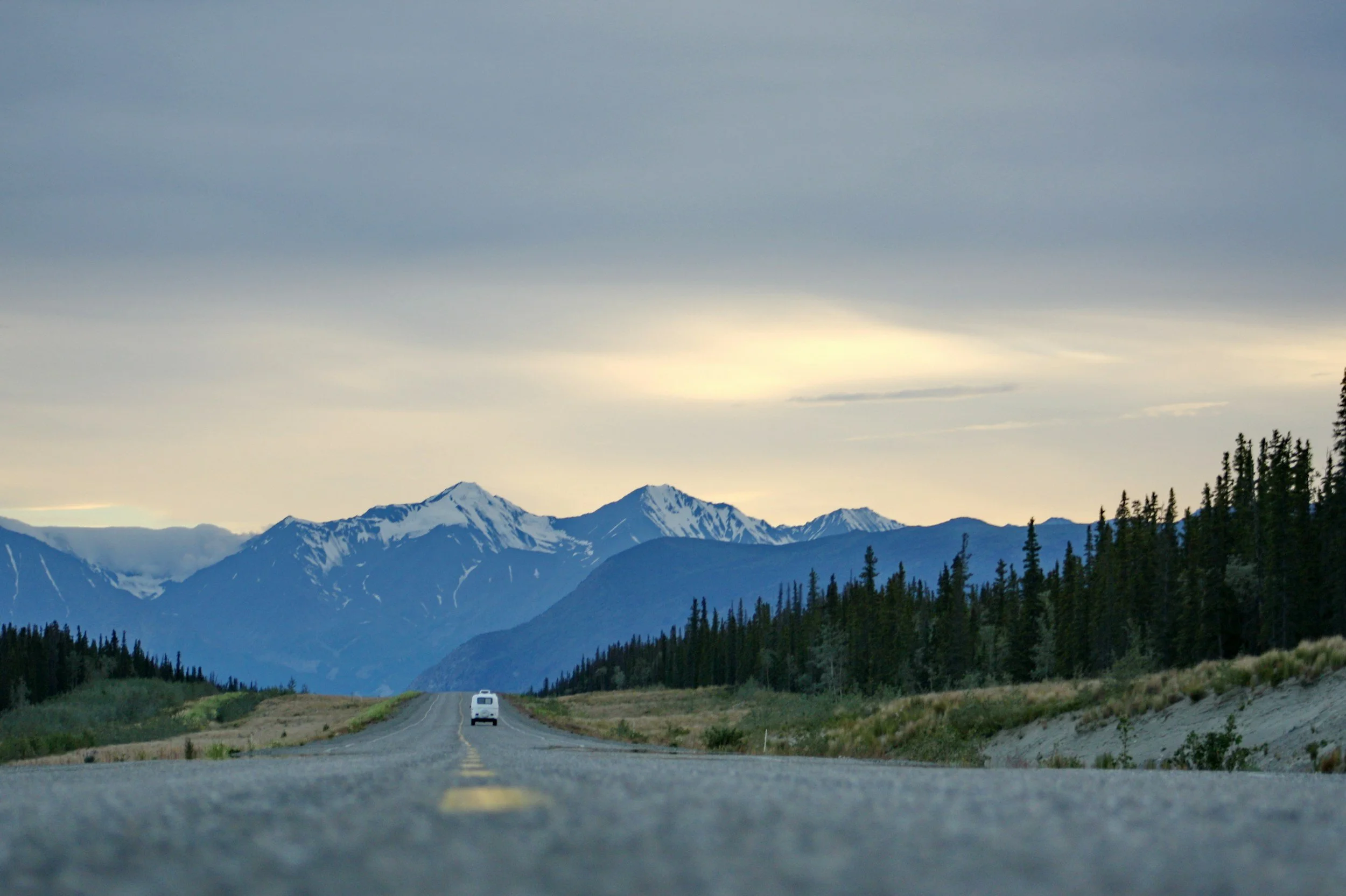 A straight road leading towards snow-capped mountains under a cloudy sky, flanked by evergreen trees on both sides.