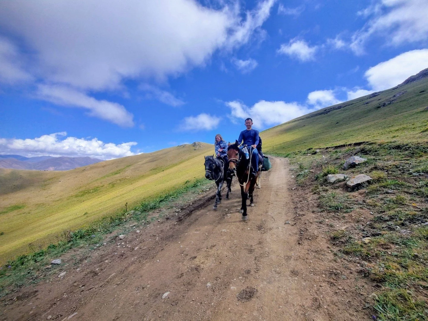 Two people riding horses on a dirt trail through a green hilly landscape under a blue sky with clouds.