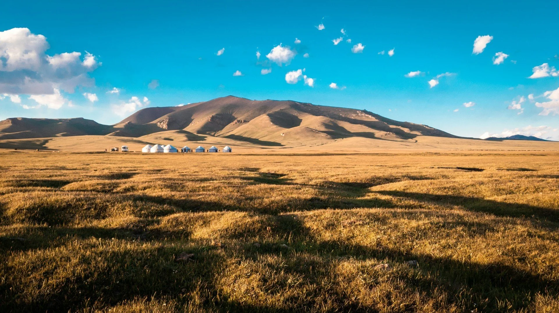 Kyrgyzstan, Central Asia. Open grassland with mountains in the background, clouds in the sky, and several white tents set up on the plains.
