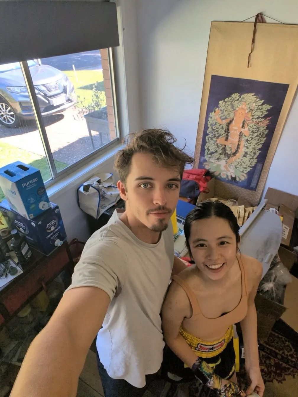 A young man with light skin, short, messy hair, and a goatee, taking a selfie with a young woman with light skin, black hair, and a big smile. They are indoors, surrounded by boxes, bottles, and miscellaneous items, with a window behind them showing a parked car outside. A large painted scroll of a deity or mythological figure hangs on the wall.