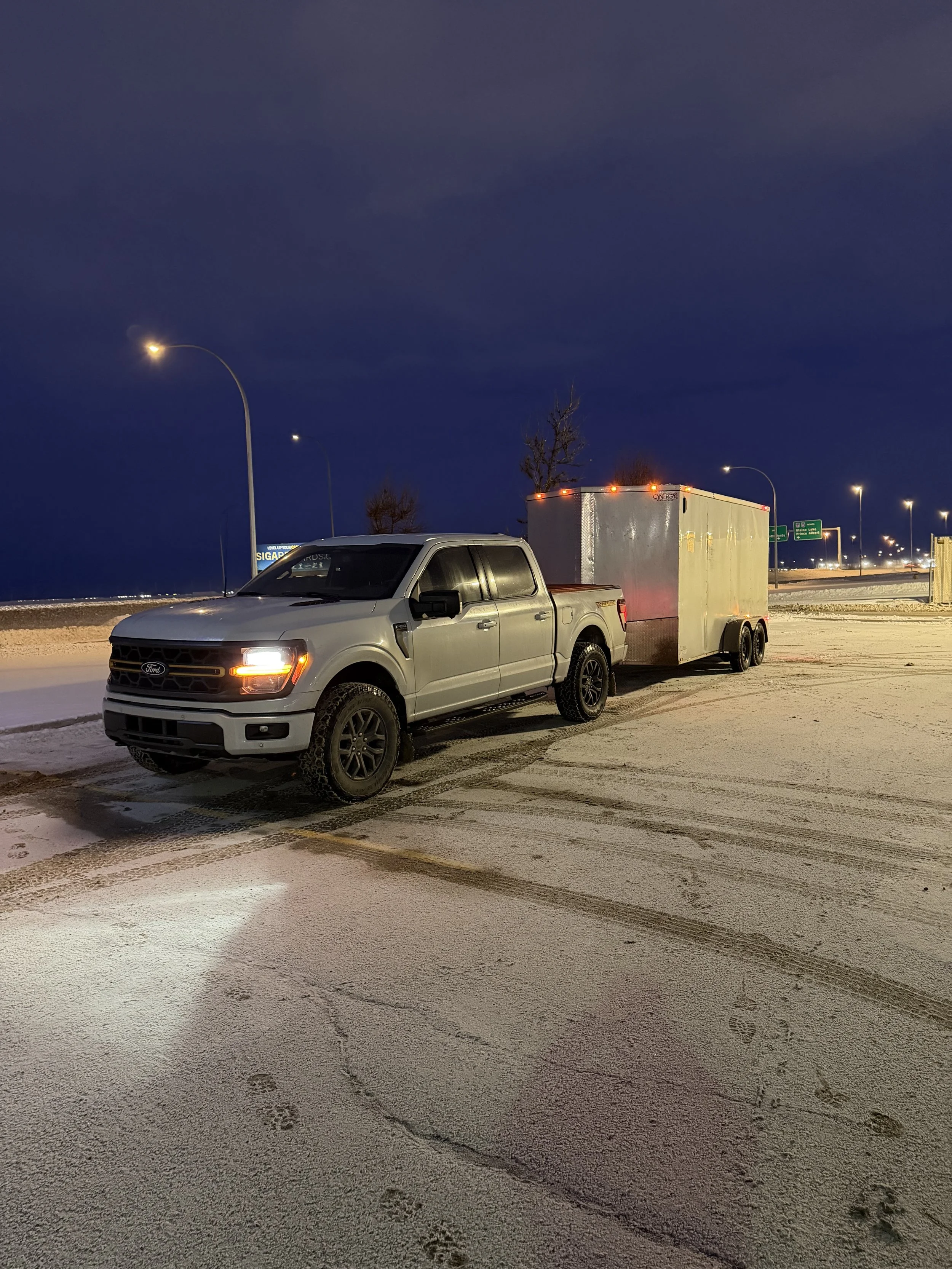 A silver pickup truck with a trailer attached, parked in a snowy parking lot at night, illuminated by streetlights.