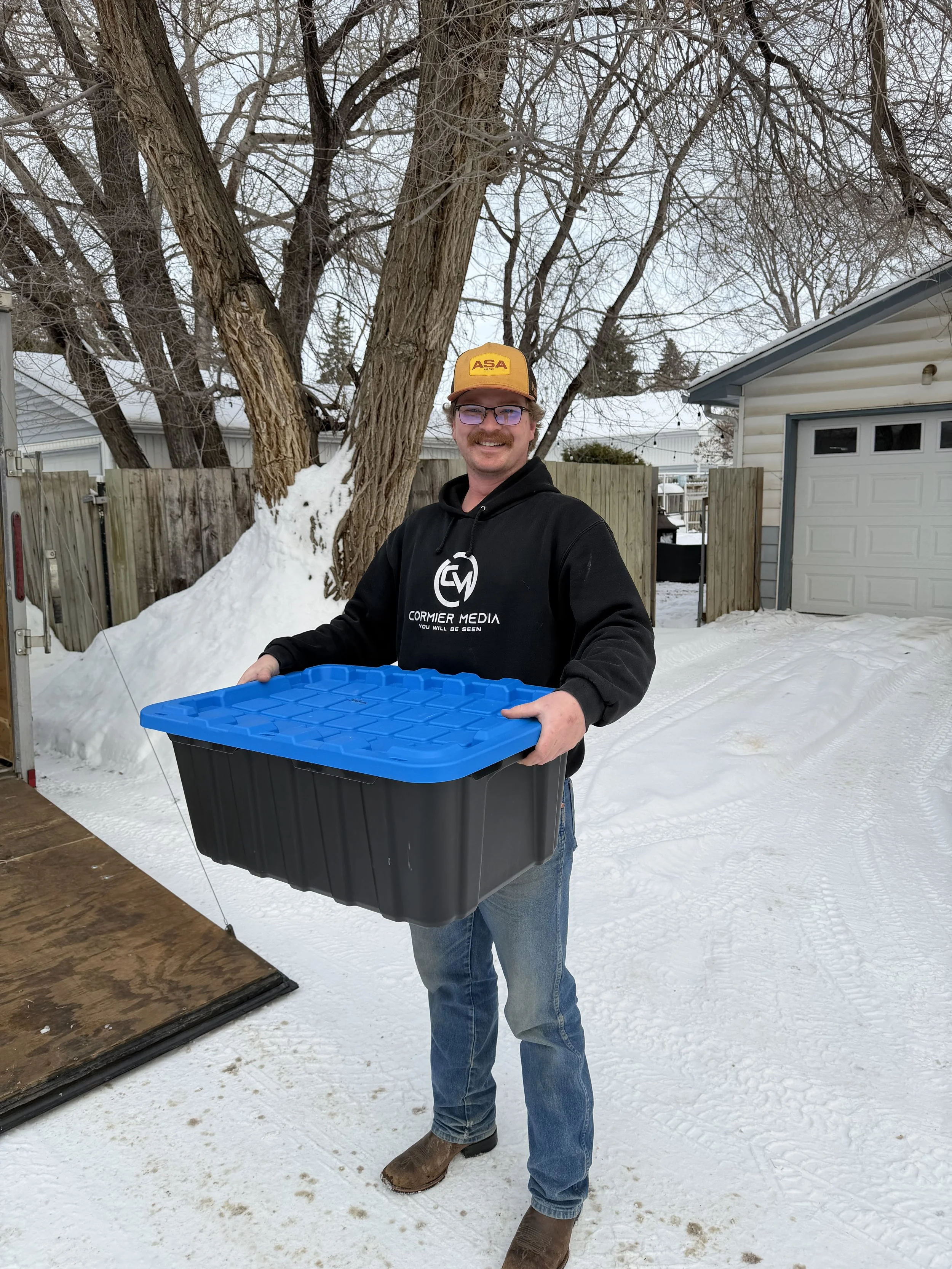 A man holding a large black plastic container with a blue lid outdoors in a snowy backyard, smiling, wearing a black hoodie, jeans, brown boots, glasses, and a yellow cap, with trees, wooden fence, and a garage in the background.
