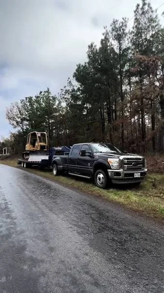 A black pickup truck parked on the side of a rural road, towing a flatbed trailer with construction equipment, trees in the background, overcast sky.