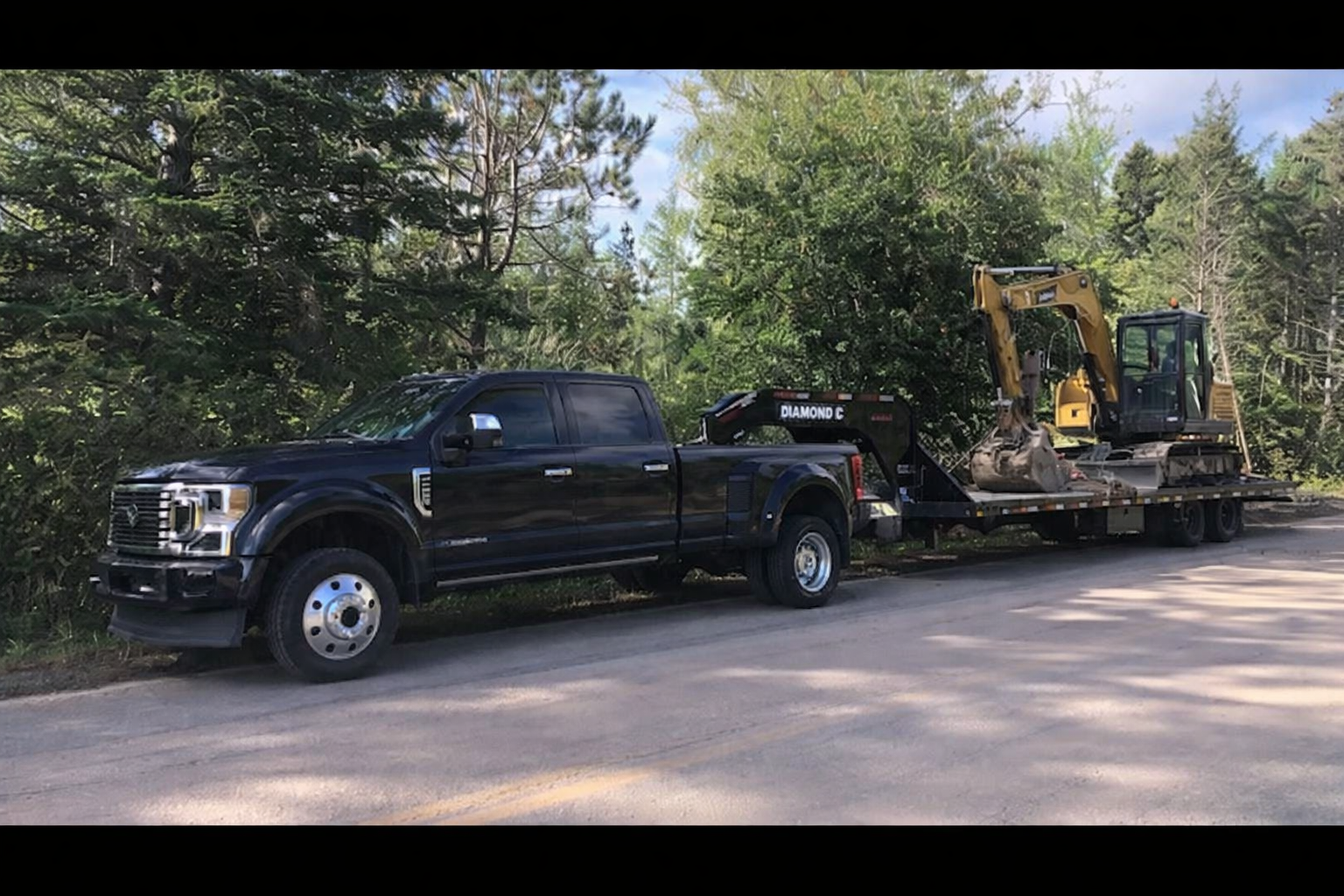 Black pickup truck towing a trailer loaded with construction equipment, including a yellow excavator, along a forested roadside.