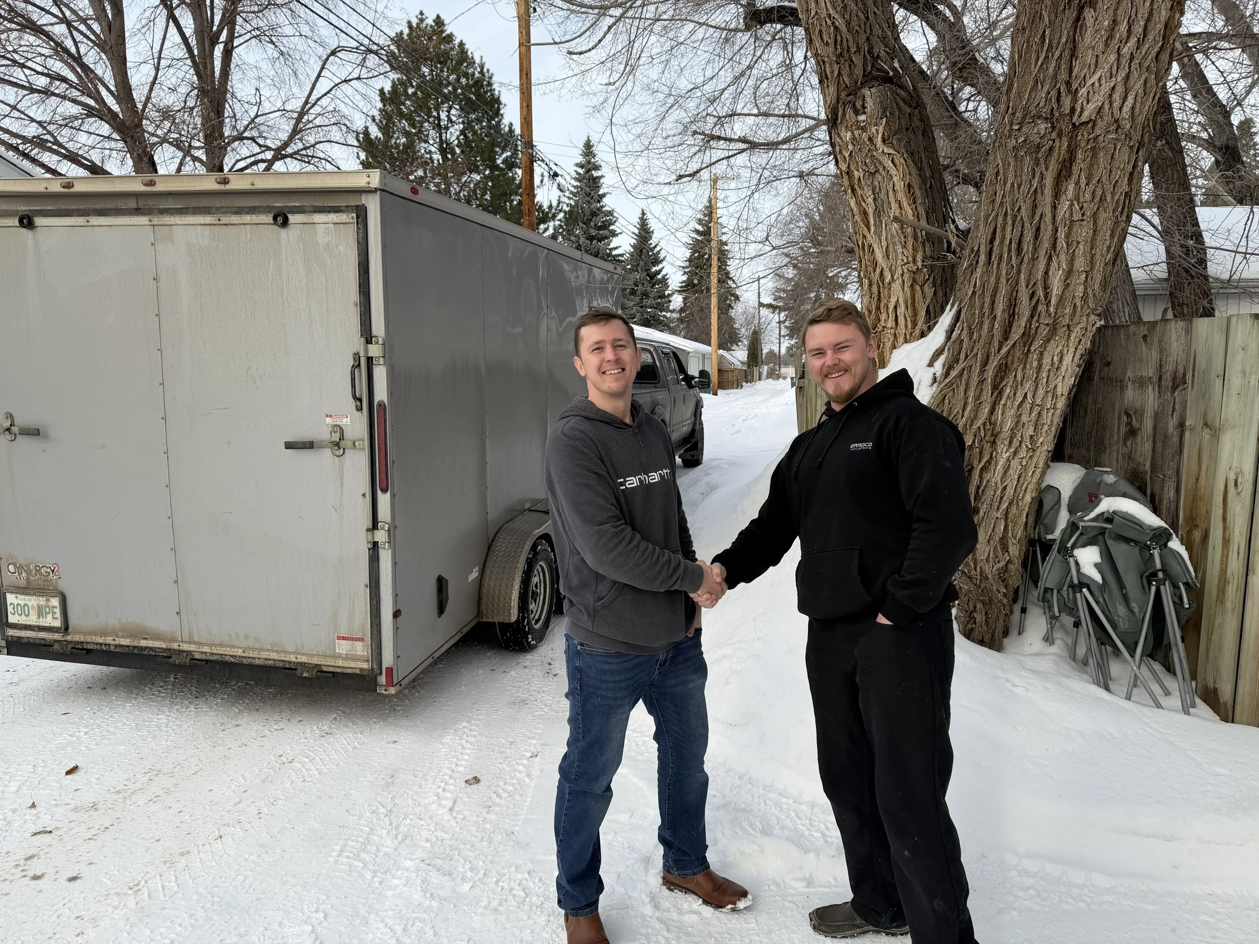 Two men in winter clothing shaking hands outdoors in a snow-covered backyard, near a large tree, with a gray enclosed trailer, a wooden fence, and snow-covered outdoor chairs in the background.