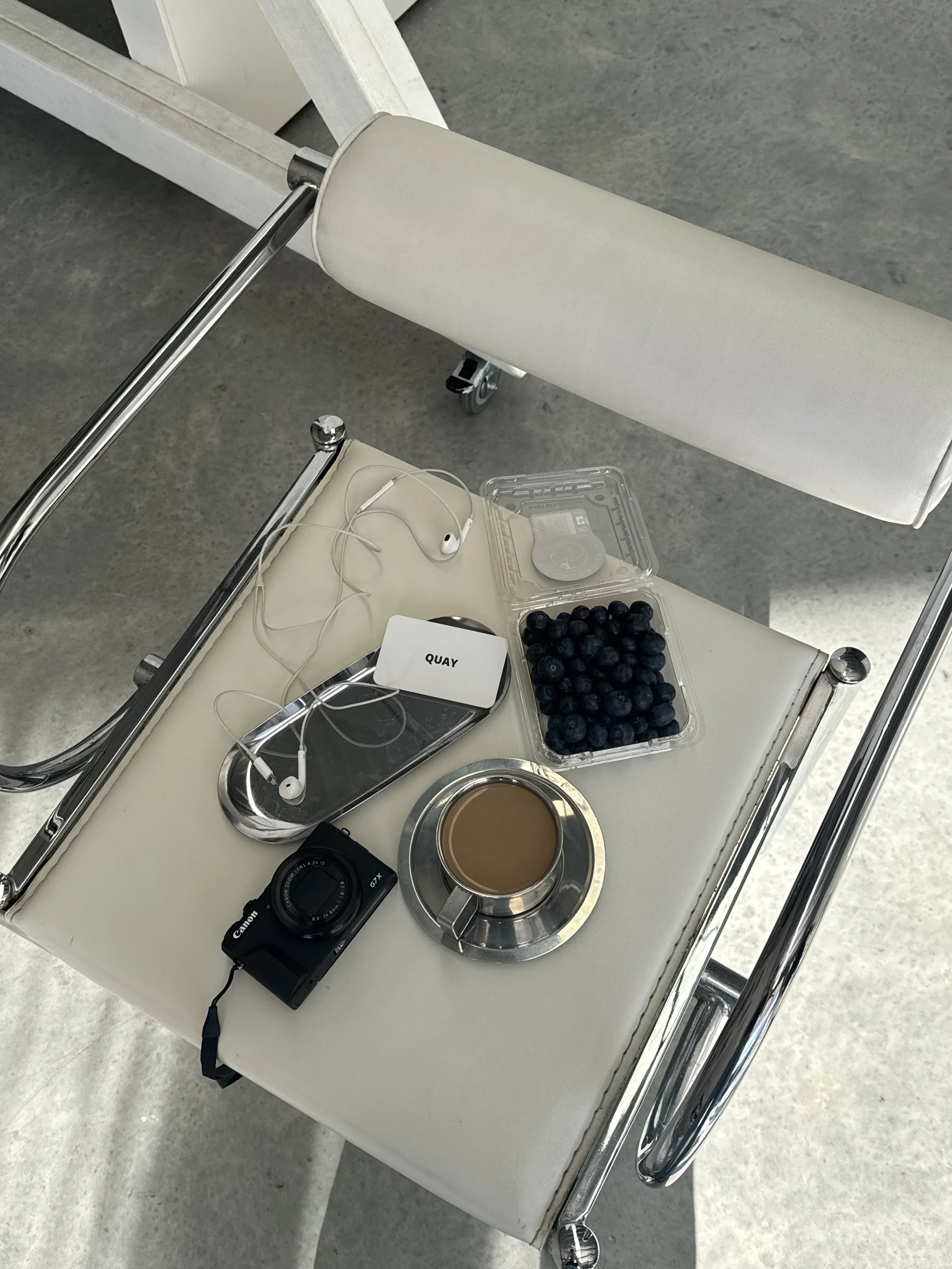 A white tray holds a black camera, a container of blueberries, a cup of coffee, a small device with earphones, and a portable charger labeled 'QUAY' inside a hospital or clinic waiting area.