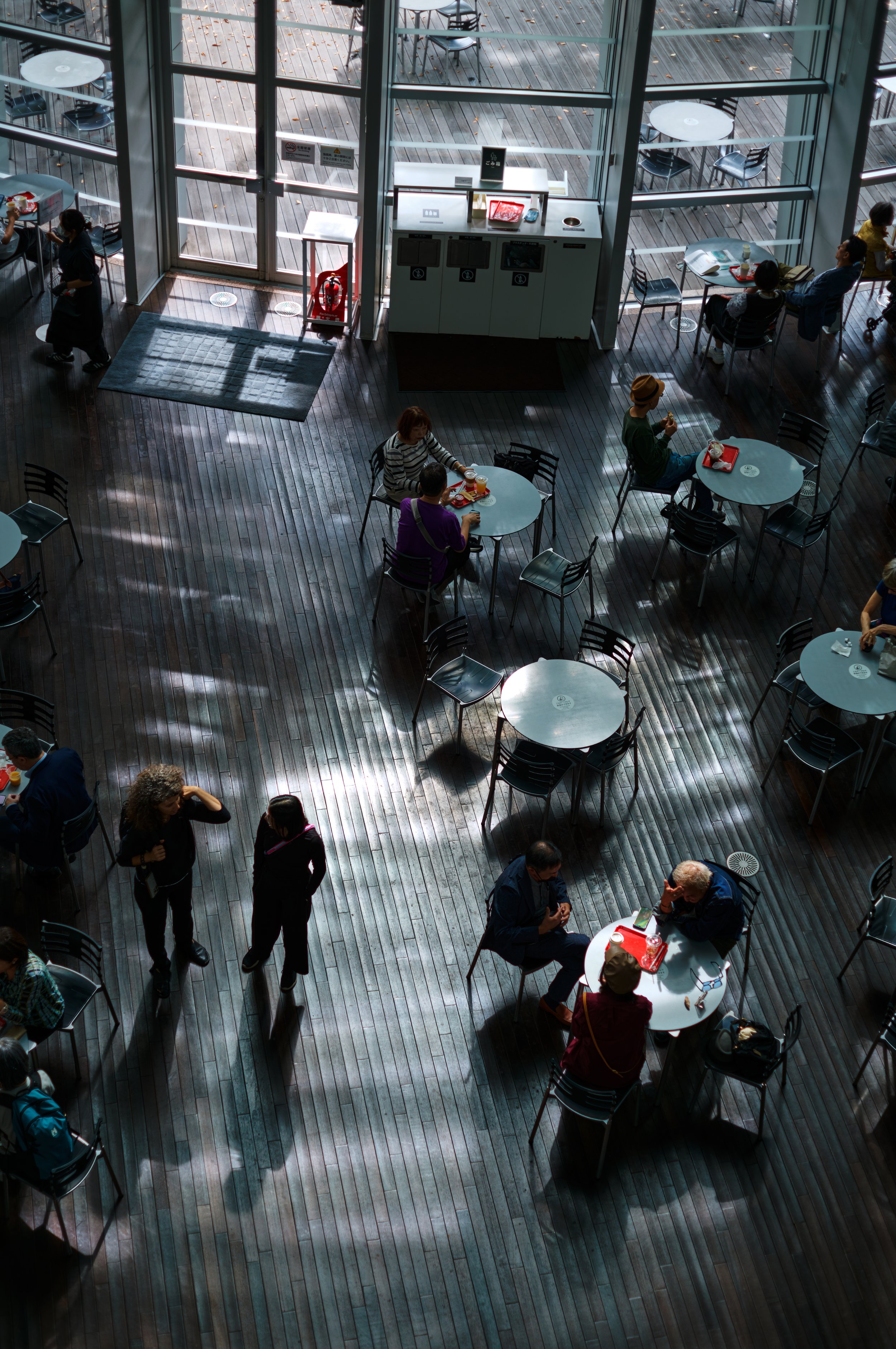 大きな窓から自然光が差し込むカフェの内部、複数の人がテーブルで食事や会話を楽しんでいます。木製の床とシンプルな家具が配置されています。
