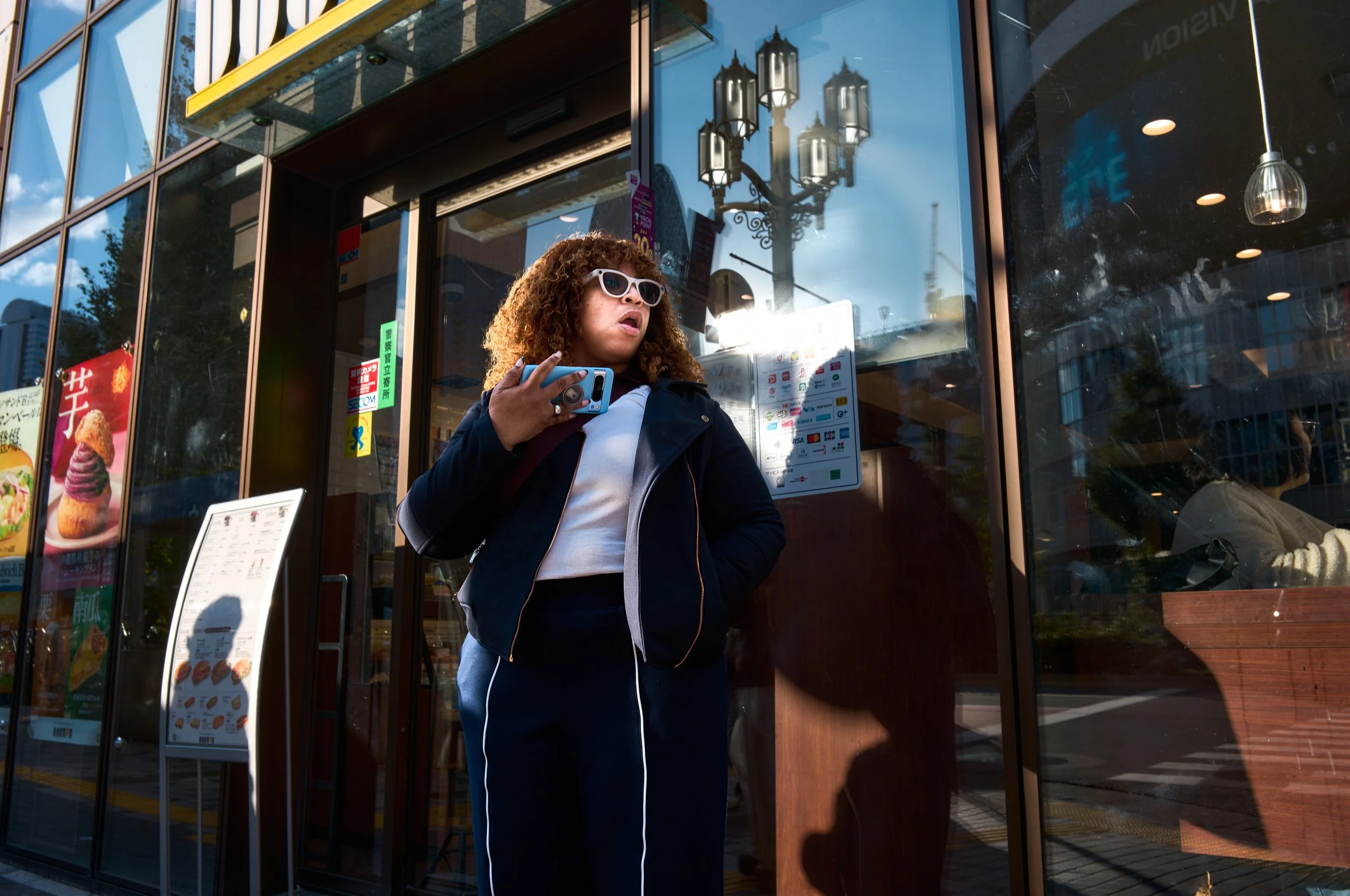 女性がスマートフォンを持ちながら外のカフェに立っている様子。背景には店内の照明や建物の反射、看板やメニューが見える。