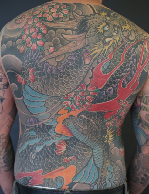 Colorful traditional Japanese-style dragon tattoo covering the person's full back to the thighs, featuring a fierce dragon with a detailed face, flowing mane, and scales, intertwined with cherry blossoms and clouds, done by tebori.