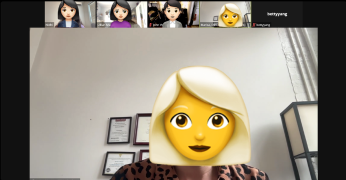 A person with a cartoon emoji face mask of a blonde woman with large eyes and dark lipstick, sitting in an office with framed certificates on the wall and a lamp to the right.