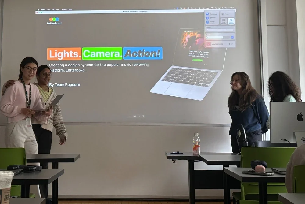 Four young women presenting in a classroom with a large projected slide about creating a design system for a movie review platform, with desks, chairs, and a computer visible.