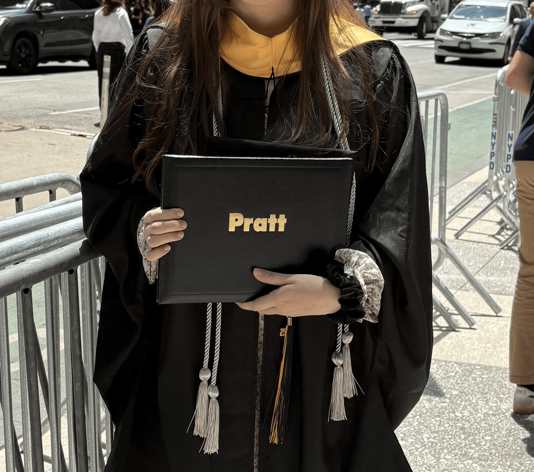 A graduate in cap and gown holding a black diploma cover with the name 'Pratt' on it, standing outdoors with other graduates and cars in the background.