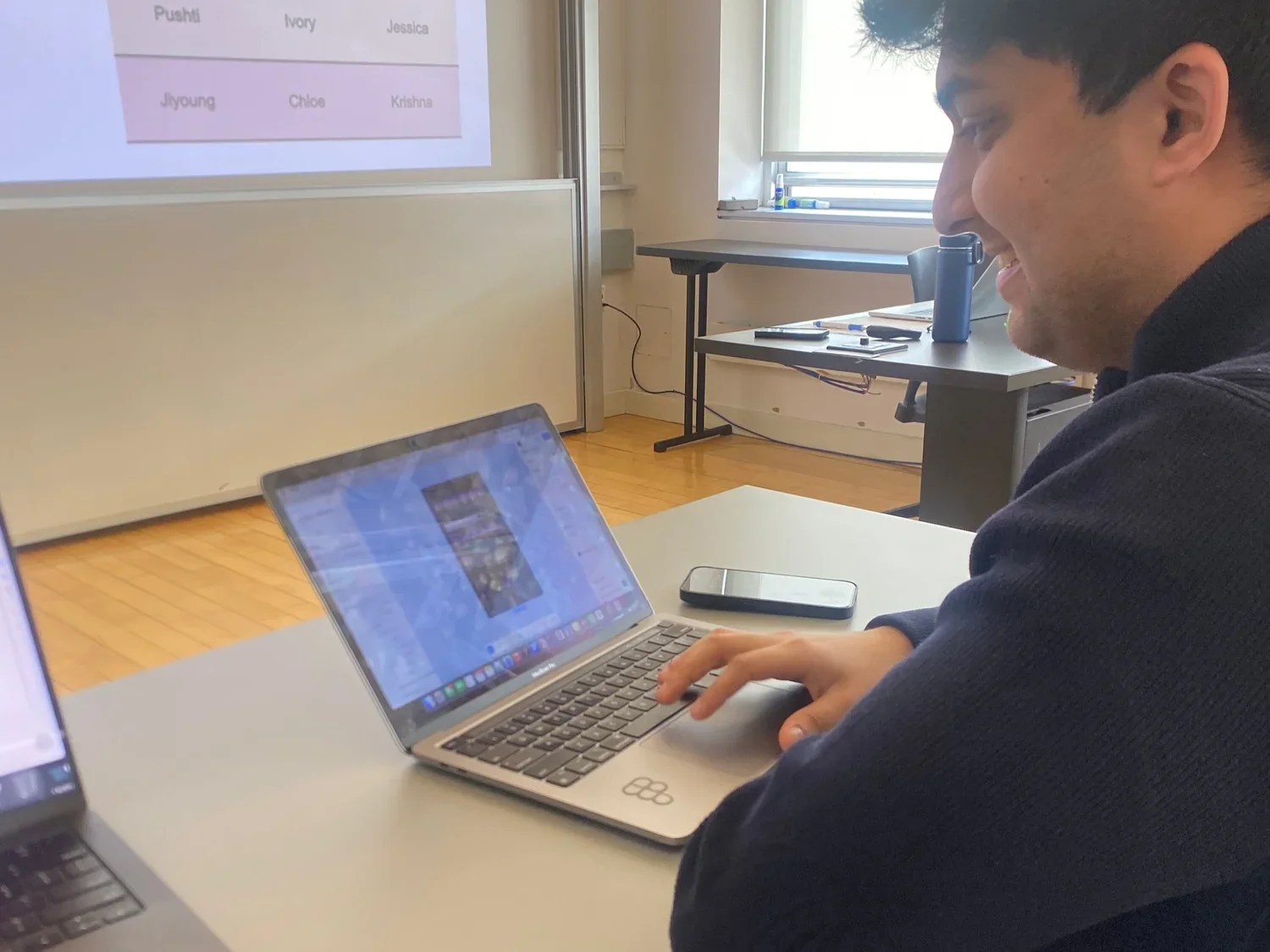 A young man sitting at a desk in a classroom, smiling while using a laptop. There is a smartphone on the desk and a window with blinds in the background.