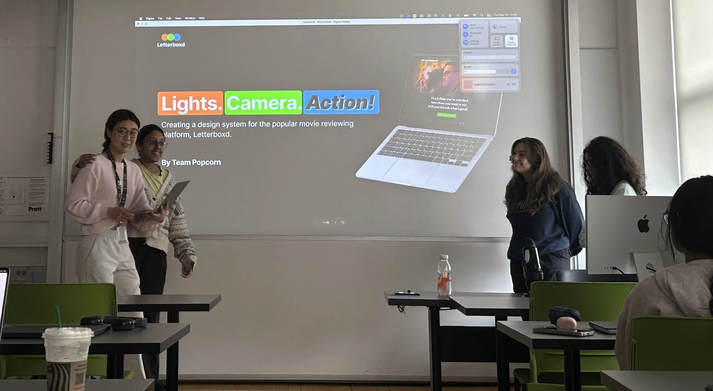 Four women presenting in a classroom, standing in front of a large screen displaying a project titled 'Lights. Camera. Action!' with text and images related to a movie review platform, and students seated at tables facing them.