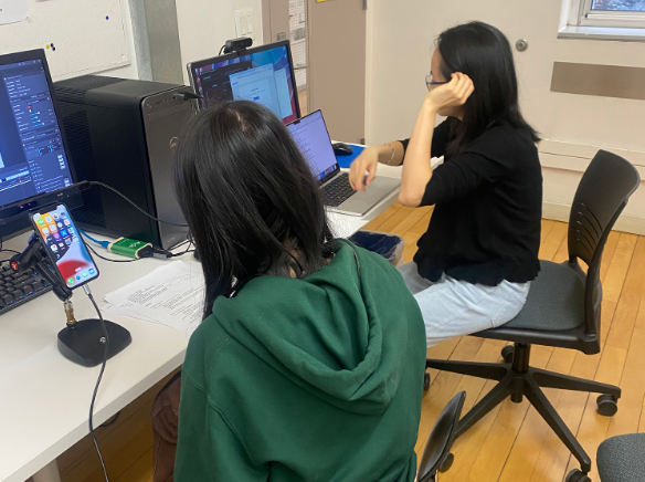 Two women working at a desk with computers, one facing away wearing a green hoodie, and the other facing sideways with glasses and black top.