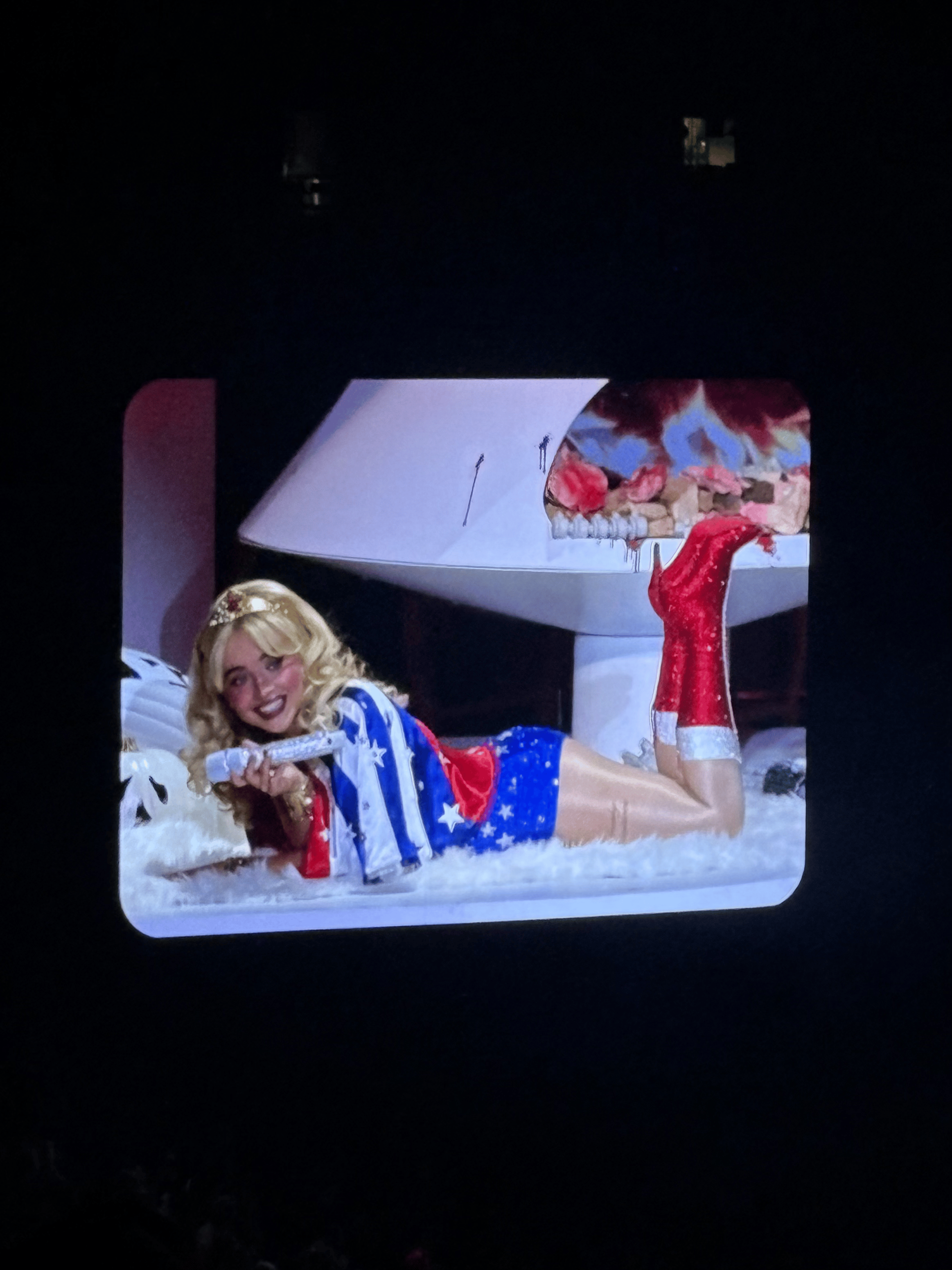 A woman lying on a fluffy surface in front of a white grand piano, dressed in patriotic red, white, and blue clothing, with red boots and a blonde wig, smiling and holding a silver object.