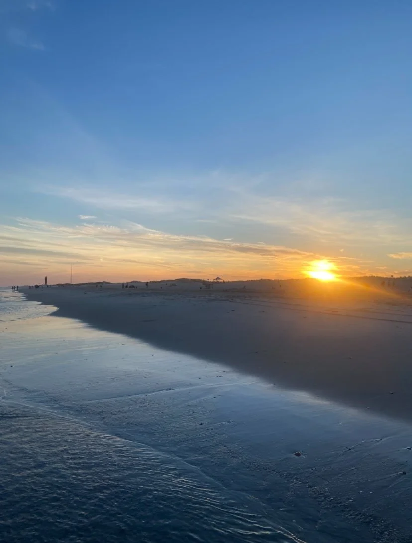 Sun setting over a sandy beach with gentle waves and a few people in the distance, under a partly cloudy blue sky.